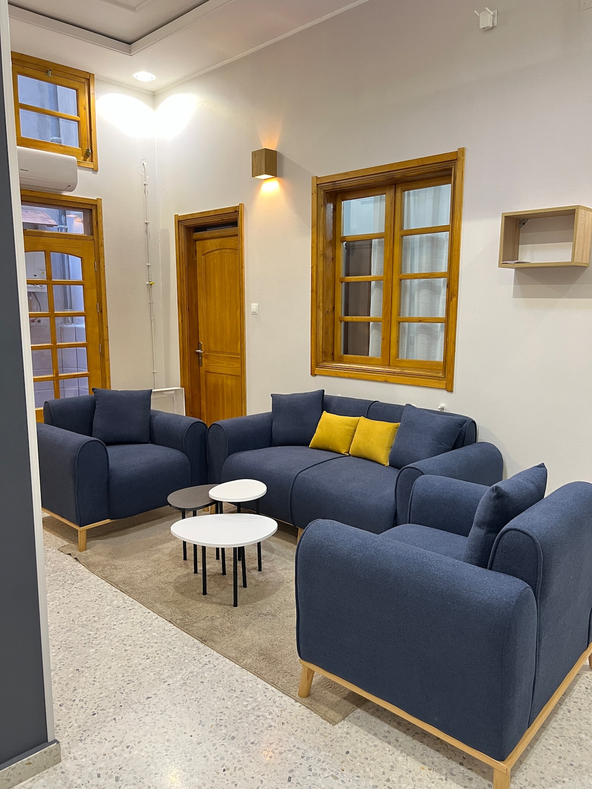 A comfortable seating area features two blue sofas and two small white tables, positioned on a neutral rug. Natural light enters through the multiple windows, highlighting the warmth of wooden doors and accents in a softly lit environment.