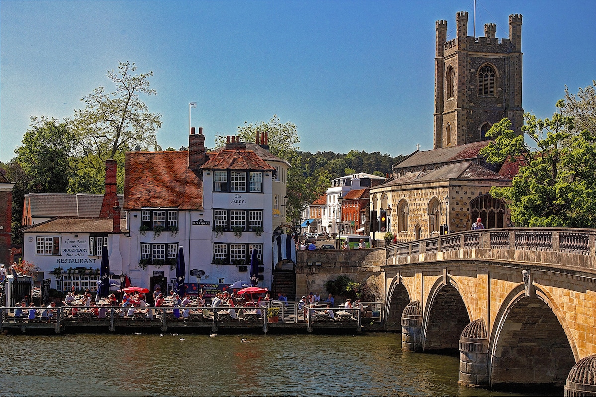 A scenic riverside view highlights charming buildings with red-tiled roofs, cafes with outdoor seating, and a stone bridge spanning the River Thames. A historic tower stands nearby, surrounded by lush greenery and a clear blue sky, reflecting the vibrancy of the Henley-on-Thames area.