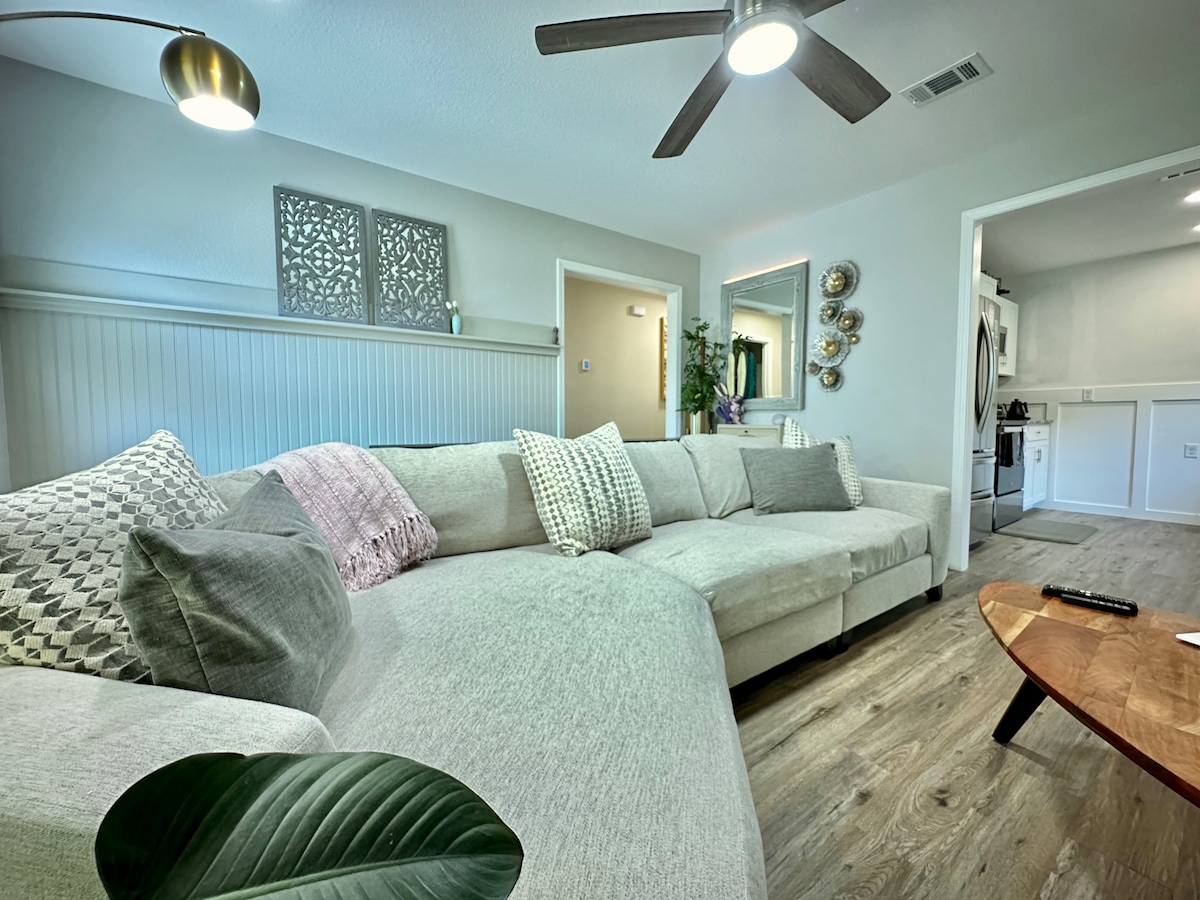 A spacious sectional sofa with various textured cushions is positioned in the cozy living area. A coffee table sits in front, and a ceiling fan circulates air above. Doors lead to a kitchen and another room, enhancing the open layout.