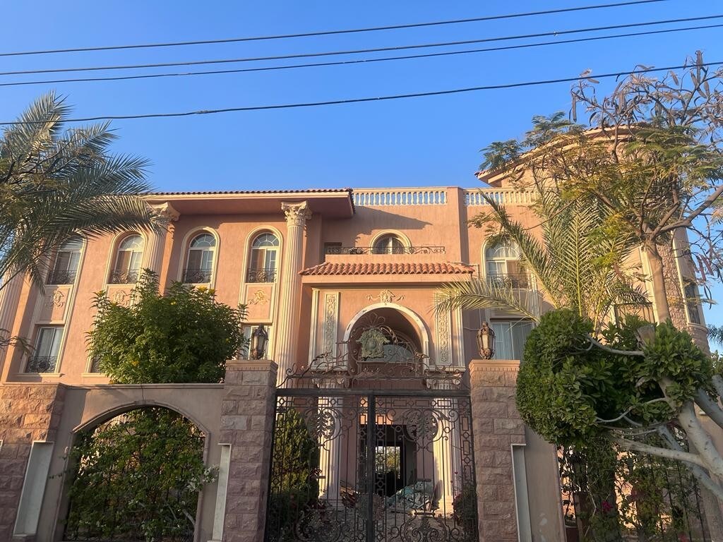 A grand villa is showcased with a distinct architectural design, featuring ornate details and multiple large windows. Lush greenery surrounds the entrance, complemented by palm trees. The facade is highlighted by a decorative gate that adds a touch of elegance to the overall appearance.