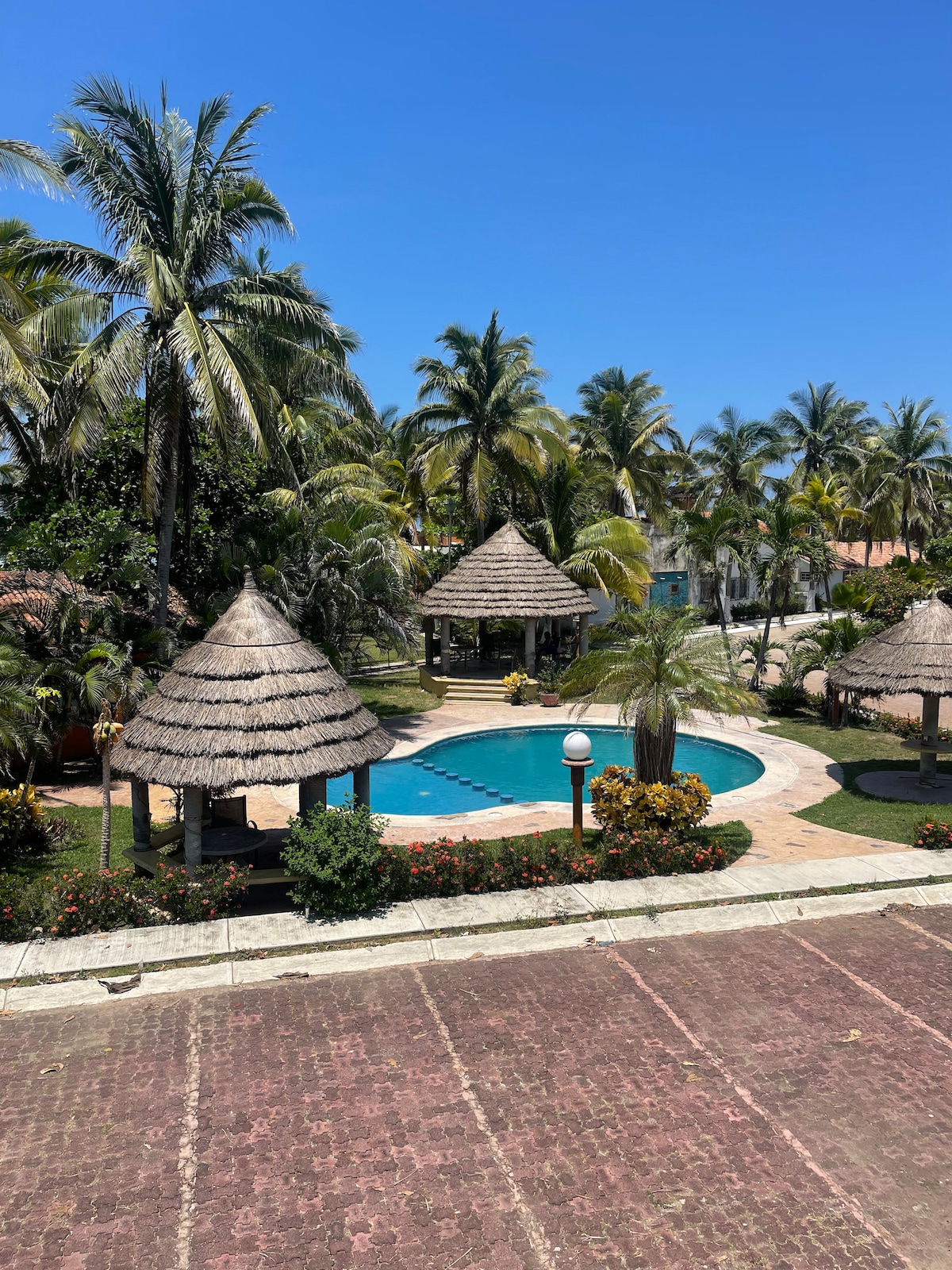 A landscaped area features a rounded swimming pool surrounded by lush palm trees and colorful flowering plants. Thatched-roof huts provide shaded seating around the pool, creating a resort-like atmosphere. A cobblestone pathway leads through the vibrant greenery, enhancing the tranquil setting.