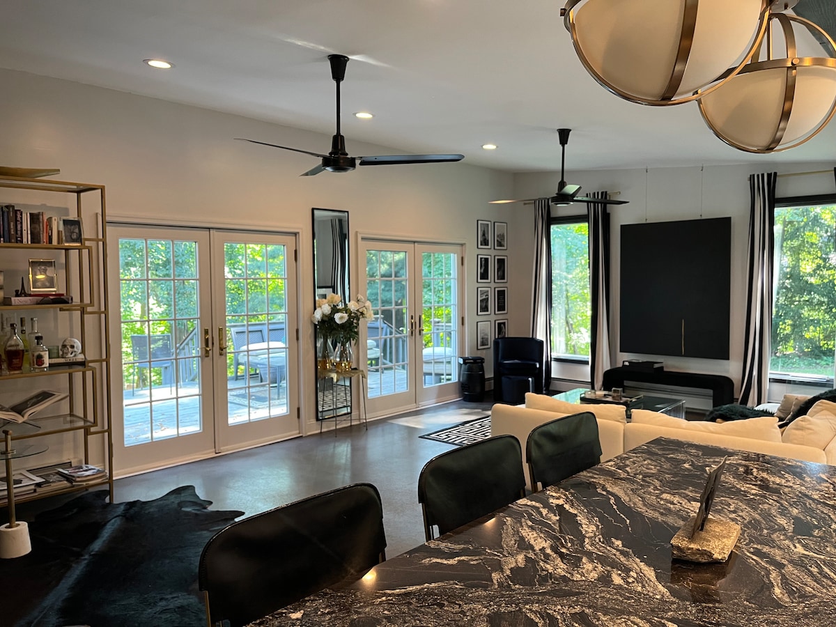 A modern living area is highlighted by large windows that fill the space with natural light, featuring comfortable seating and stylish decor. A sleek marble countertop is visible in the foreground, and a curated bookcase adds to the artistic ambiance. Ceiling fans enhance airflow.