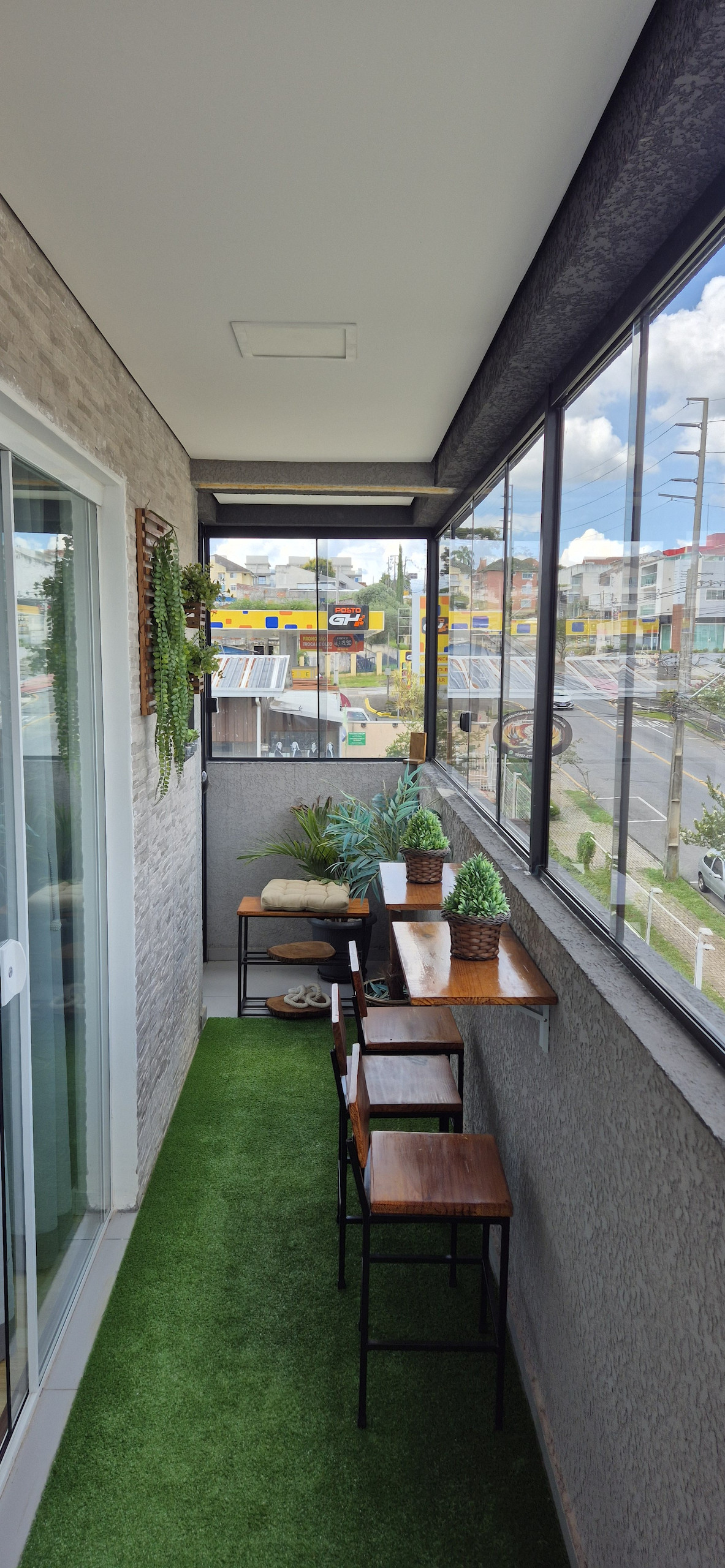 A balcony is presented, featuring glass railings that offer a clear view of the surroundings. Potted plants and decorative items are placed on a tabletop. Two wooden chairs sit alongside a small table on a green carpet, creating a comfortable outdoor seating area.