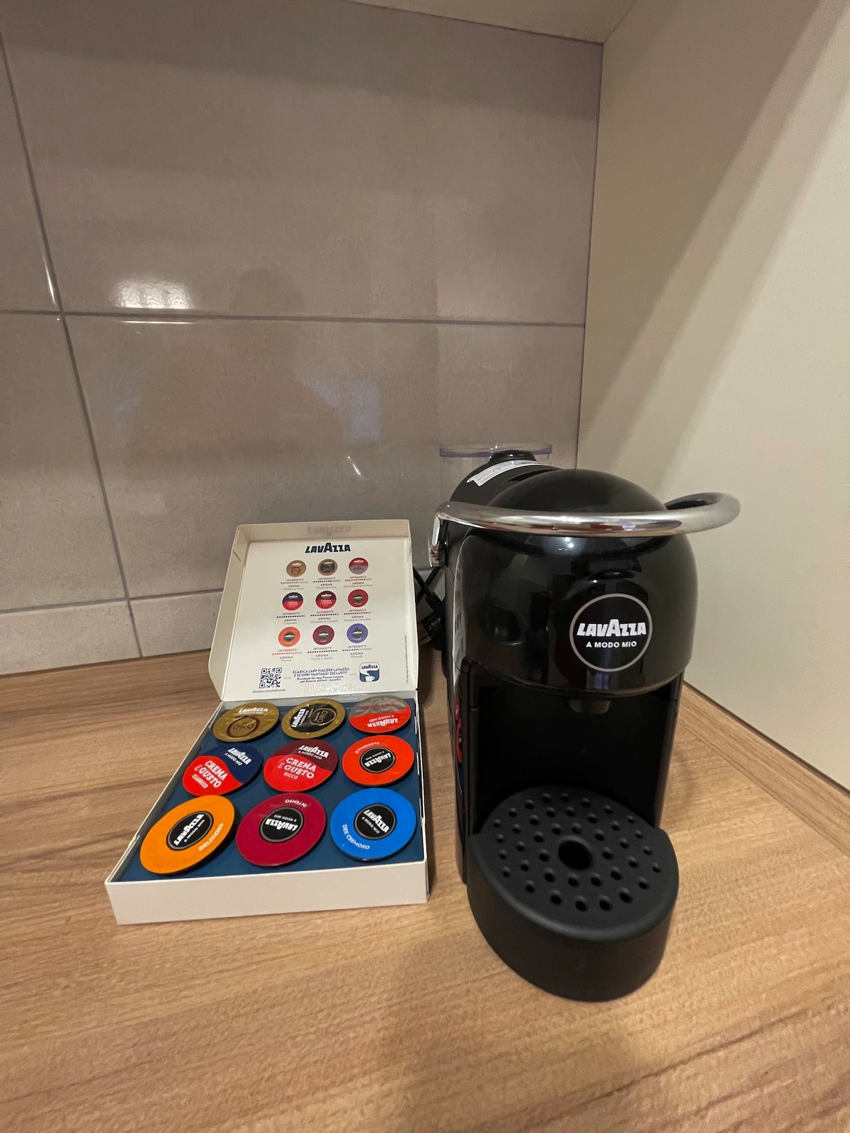 A sleek coffee machine is displayed on a wooden countertop, accompanied by a box of assorted coffee pods in various colors. The machine's design features a polished black finish with a prominent brand logo, creating a modern and functional coffee station.