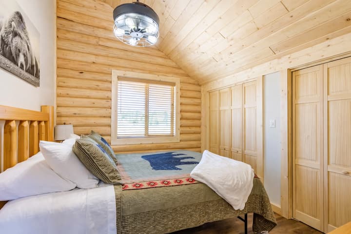 A warm, cabin-style bedroom with a king bed, simple wood furnishings, and soft lighting. The room includes a wall-mounted TV, closet space, and easy access to the loft area
