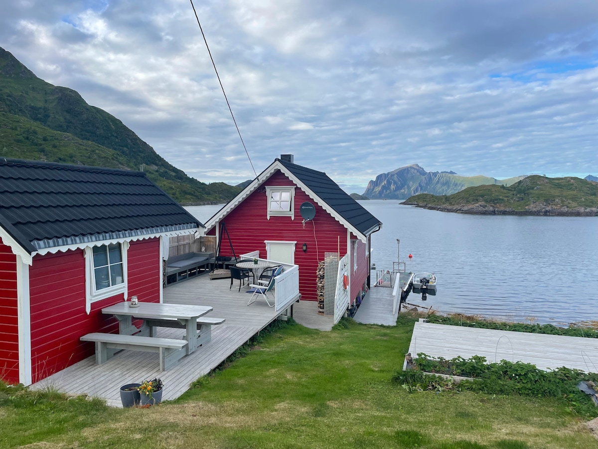 A charming red cottage sits on a grassy slope, overlooking the calm waters. A wooden deck features outdoor seating and a dining table. Mountains rise in the background against a cloudy sky, while a small boat can be seen docked nearby.