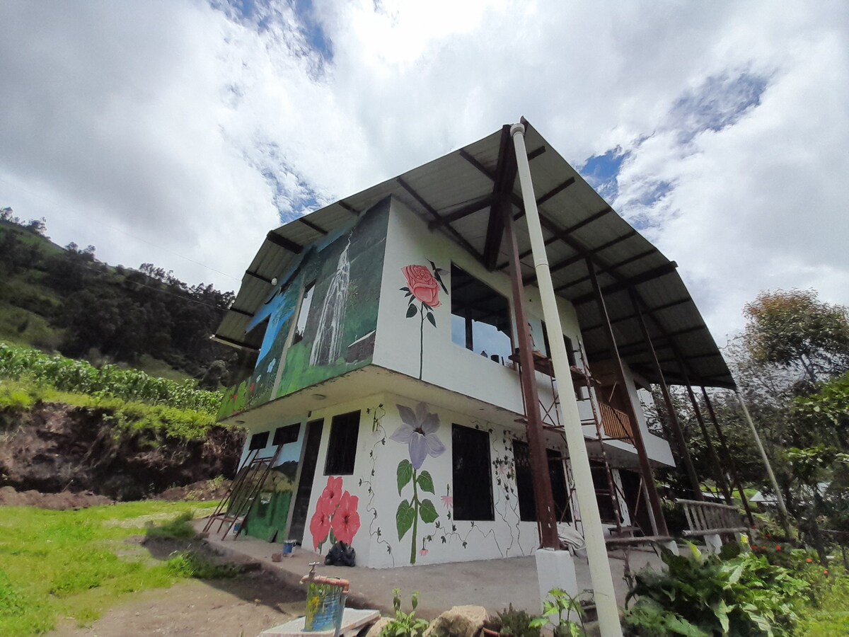 A two-story building is depicted with vibrant murals showcasing various flowers on its exterior. Large windows offer views of the surrounding greenery, while the structure is supported by metal beams. A blue planter sits at the base, blending harmoniously with the natural environment.
