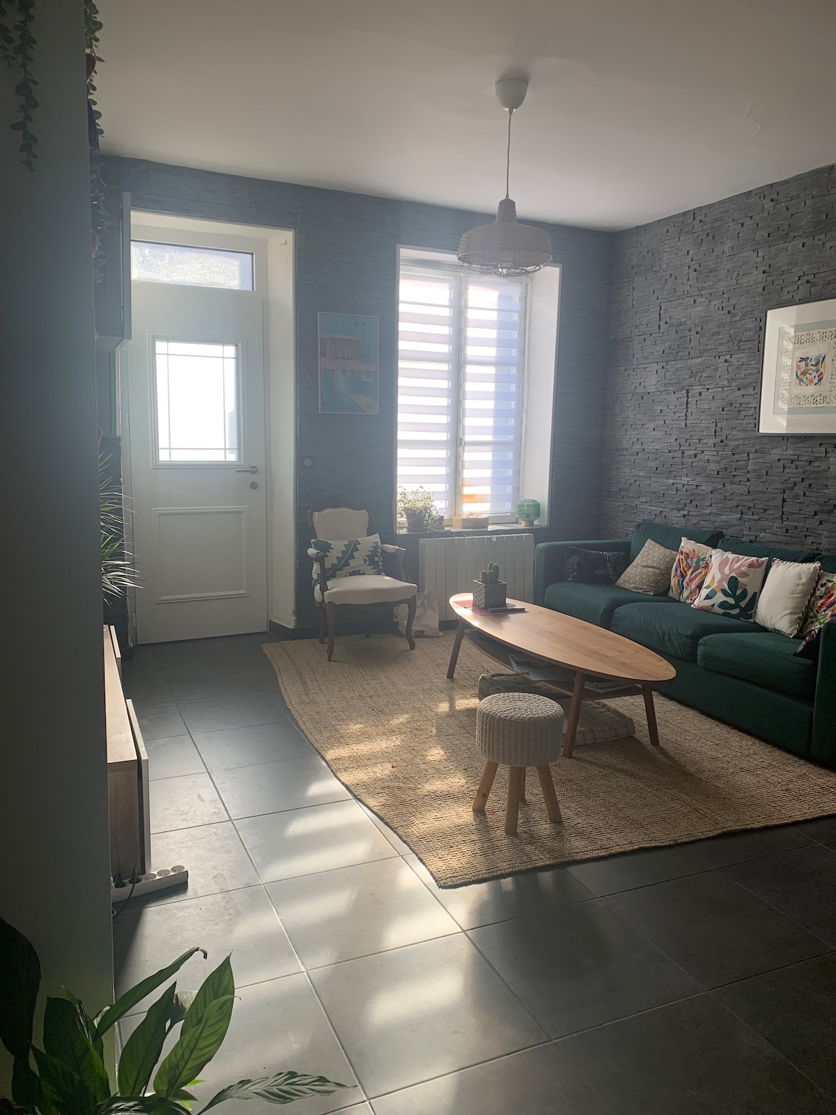 A light-filled living area features a dark stone wall and a cozy green sofa adorned with patterned cushions. Natural light streams through a window covered by horizontal blinds, illuminating a wooden coffee table and a textured area rug. A small stool adds additional seating.