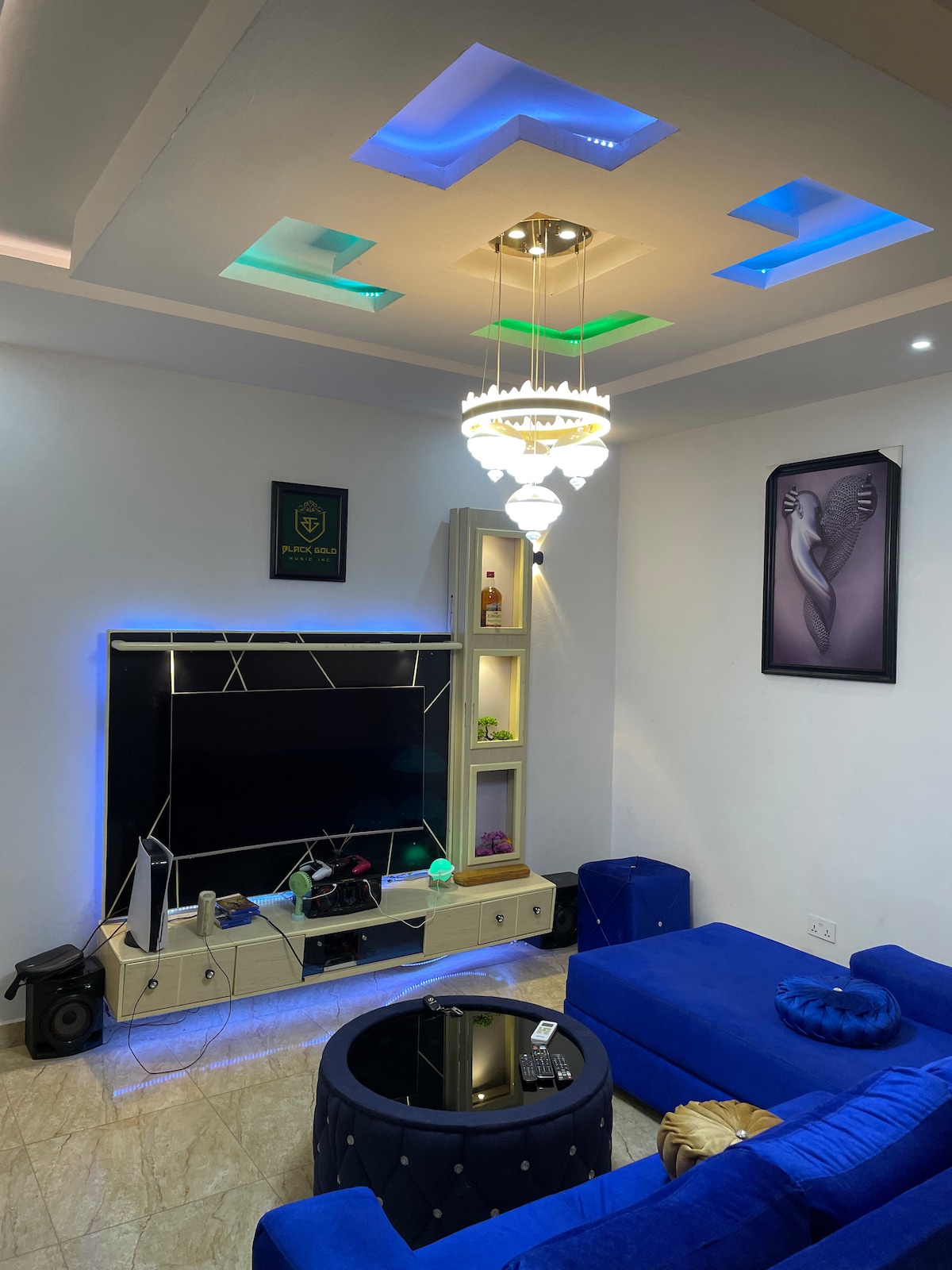 A modern living area features a blue sectional sofa arranged around a round coffee table. A flat-screen TV is mounted against a stylish wall unit, illuminated by ambient lighting. Decorative shelves display various items, and a unique chandelier adds visual interest above.