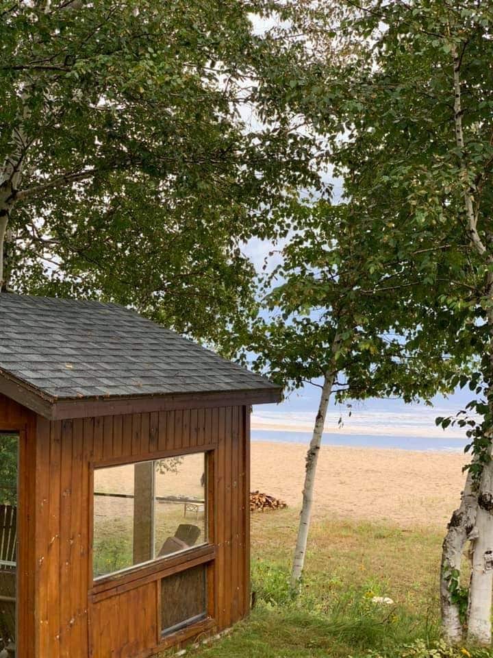 A rustic wooden cabin is framed by leafy trees, offering a glimpse of the sandy beach beyond. Several outdoor chairs are visible on the cabin's porch, providing a comfortable space to enjoy the tranquil surroundings.