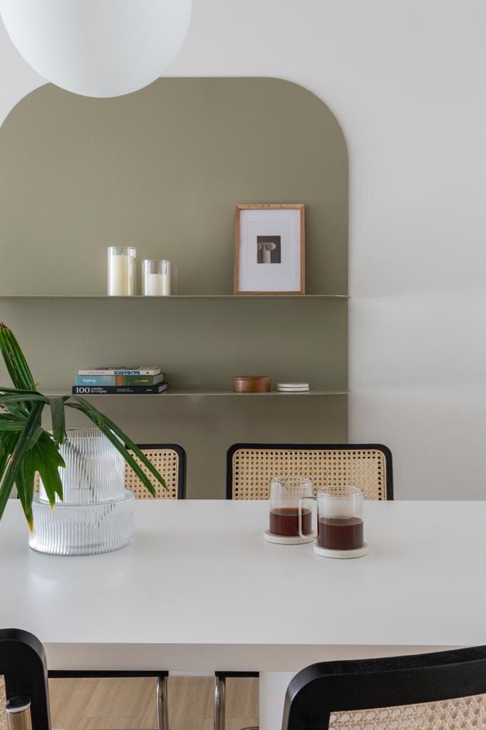 A modern dining area features a white table surrounded by dark chairs with woven backs. A green archway shelf displays books, a framed picture, and candles, while a glass vase with foliage adds a natural element to the space.