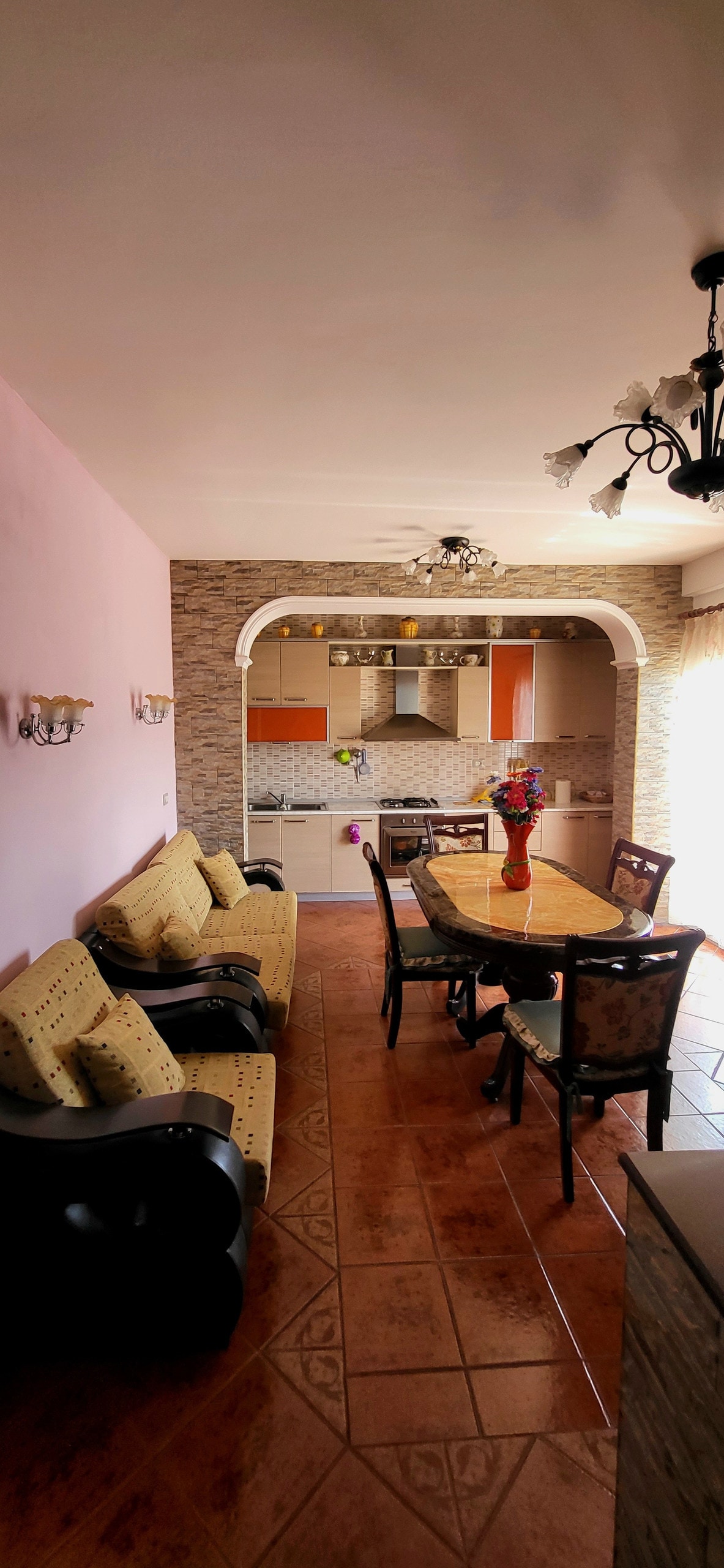 A communal dining area is featured, showcasing a wooden table surrounded by several chairs. The kitchen is visible in the background, equipped with modern appliances. Comfortable seating is arranged near the table, and soft lighting enhances the welcoming environment.