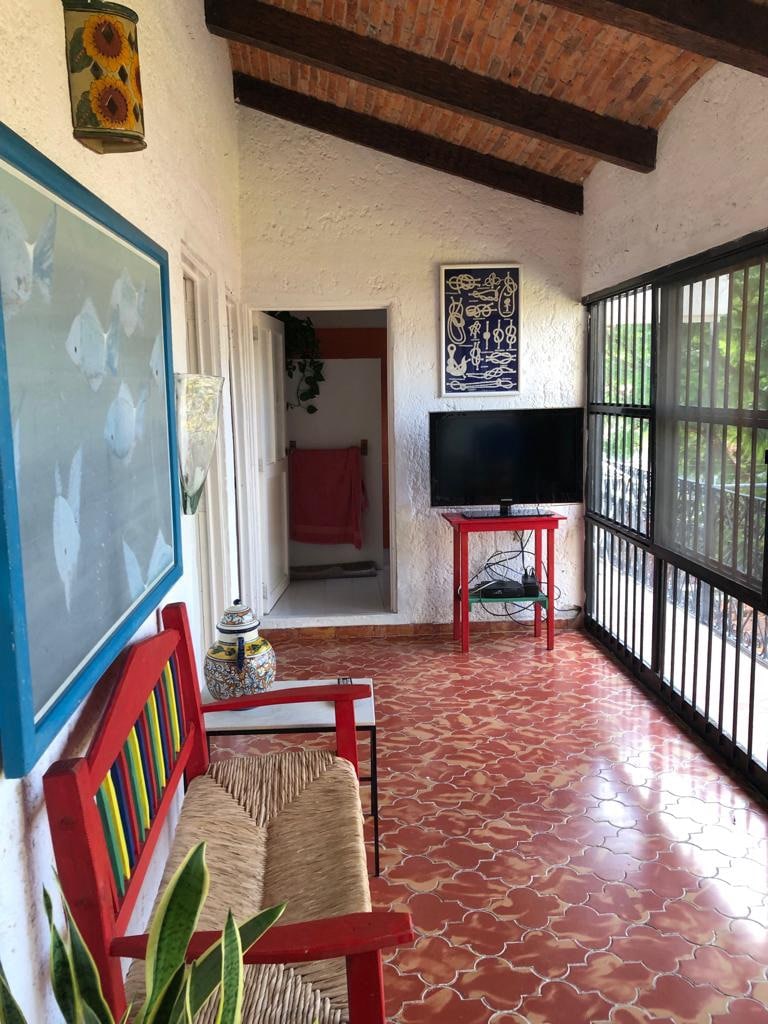 A sunlit corridor features a colorful bench and woven chair, complemented by tropical plants. The tiled floor reflects the natural light. Artwork adorns the walls, and a television is positioned on a vibrant red stand. Windows provide a view of the outdoors while ensuring a spacious feel.