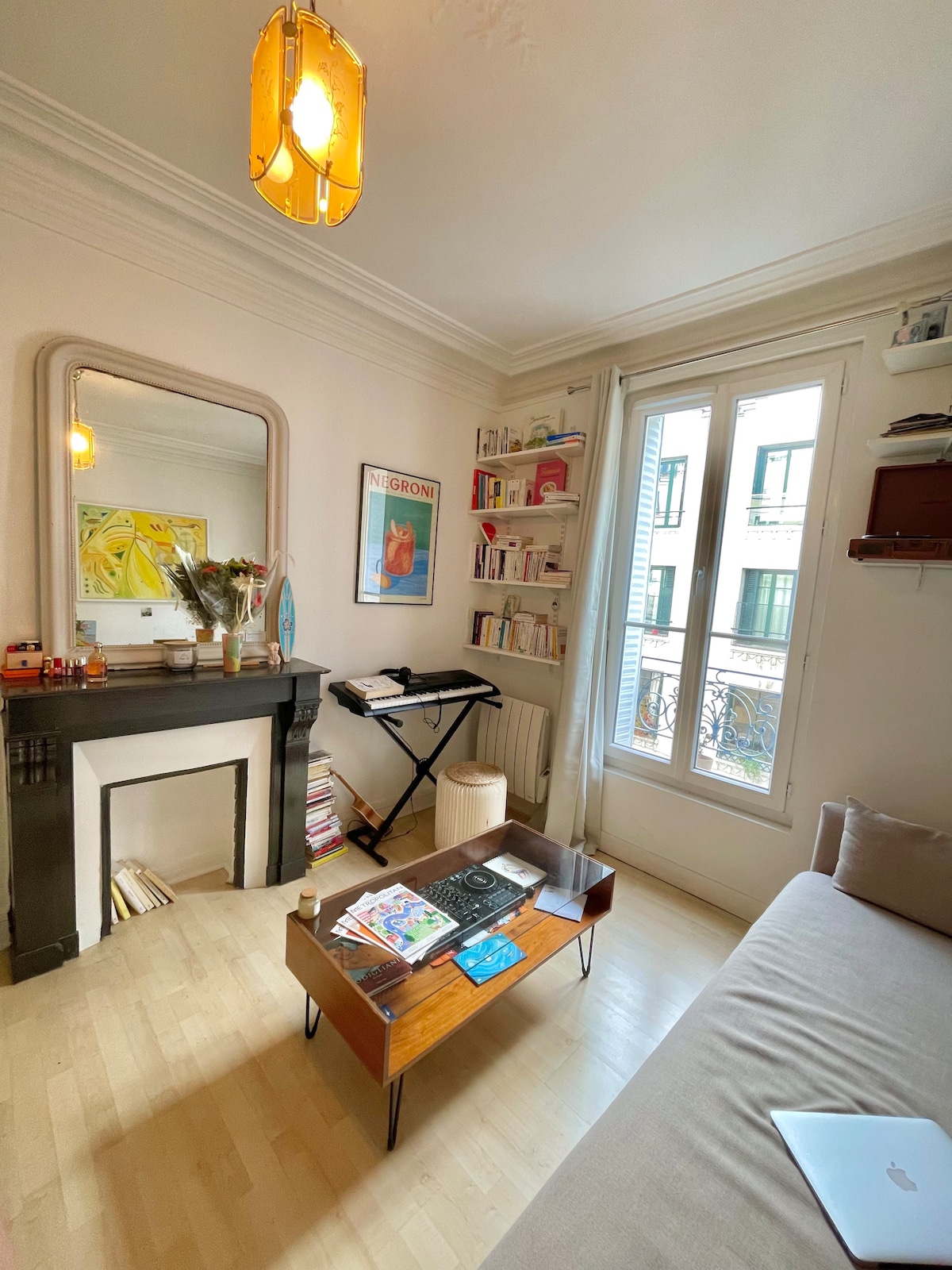 A living area features a light-colored sofa opposite a wooden coffee table, adorned with magazines and decorative items. A black fireplace adds character alongside a mirror reflecting the space. Natural light enters through a set of tall windows, flanked by bookshelves filled with various books.