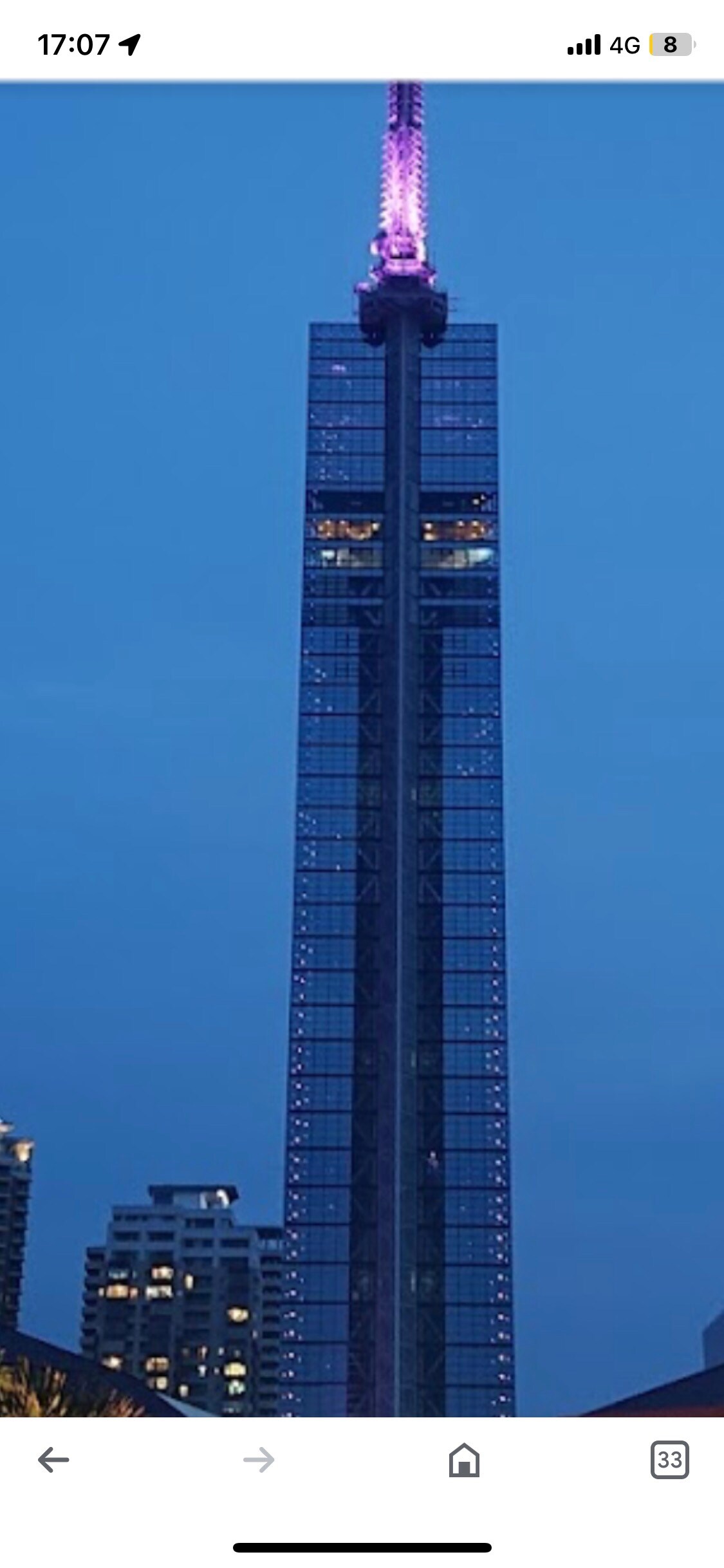 A tall building with a sleek glass facade is illuminated in purple light against a twilight sky. The structure features a prominent spire atop, and surrounding buildings are visible at the base, creating a city skyline view.
