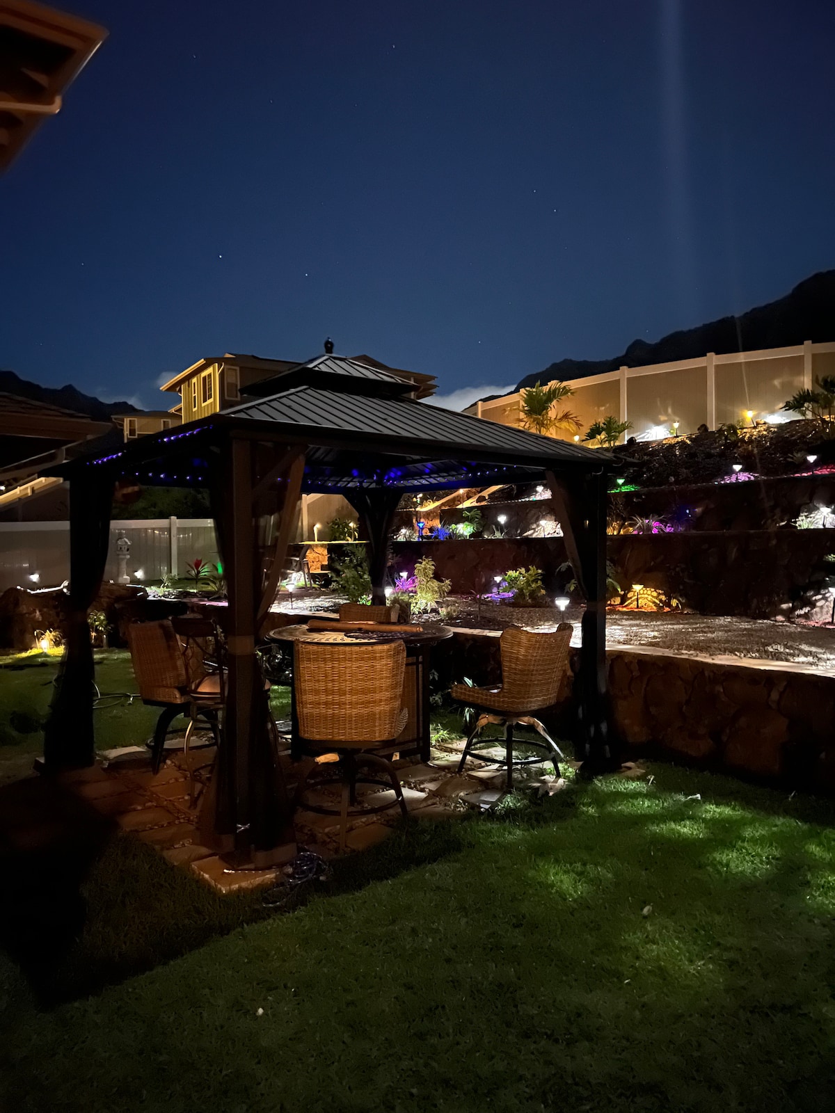 A serene outdoor gazebo is illuminated at night, surrounded by landscaped gardens. Four wicker chairs are arranged around a circular table, creating an inviting space for relaxation. Soft outdoor lighting highlights the greenery and mountain backdrop, enhancing the tranquil atmosphere.