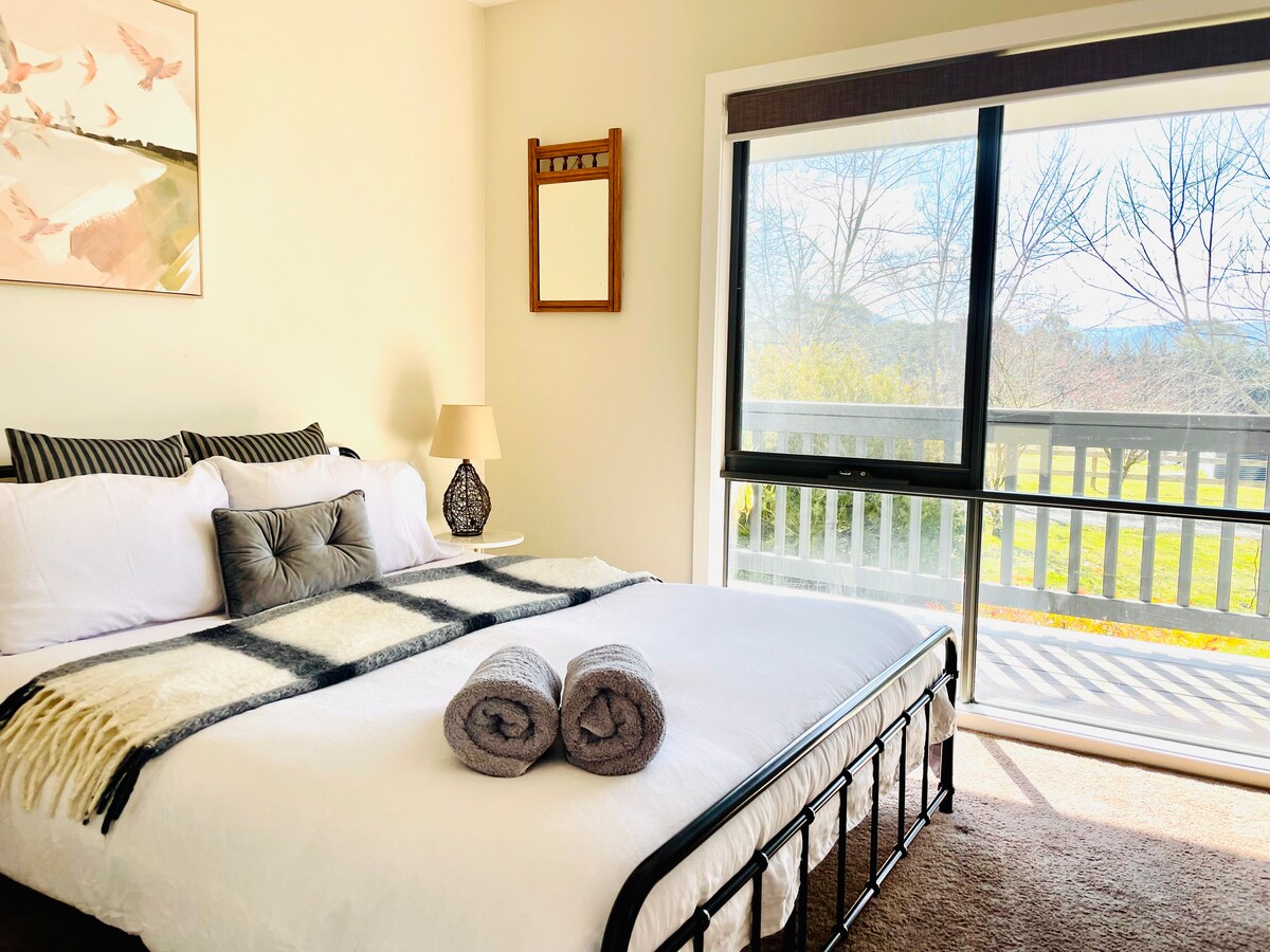 A well-lit bedroom features a spacious bed dressed in white linens and a patterned throw. Two rolled towels are placed neatly at the foot of the bed. Large glass doors offer views of the garden and mountains, allowing natural light to fill the room.