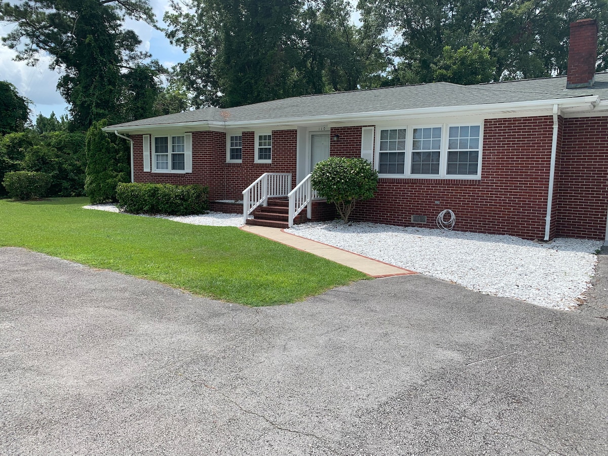The brick ranch home is set on a grassy lawn, featuring a concrete walkway leading to a white front porch with steps. Gravel borders the pathway, and well-maintained greenery adds to the inviting entrance.