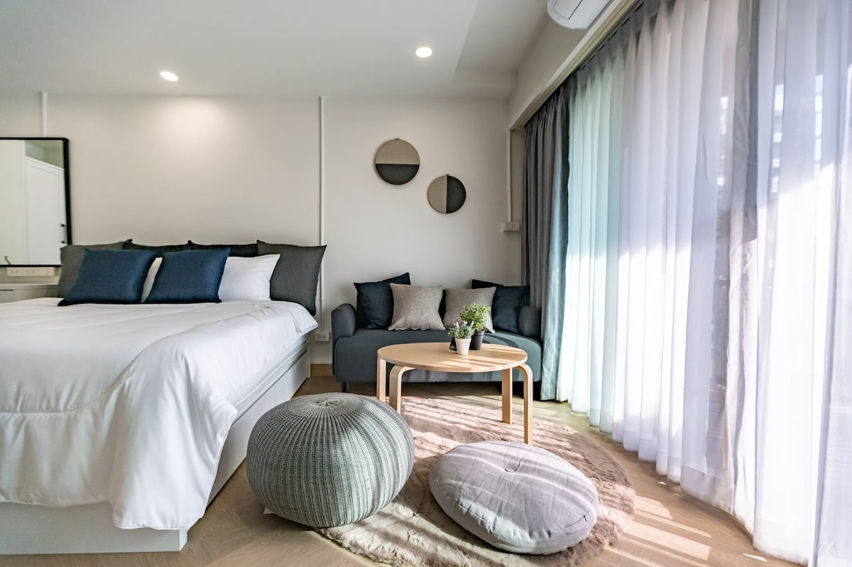 A modern room features a spacious bed with neatly arranged pillows, complemented by a cozy seating area with a small sofa. A minimalist coffee table is surrounded by round cushions on a textured rug. Natural light filters in through large, sheer curtains.