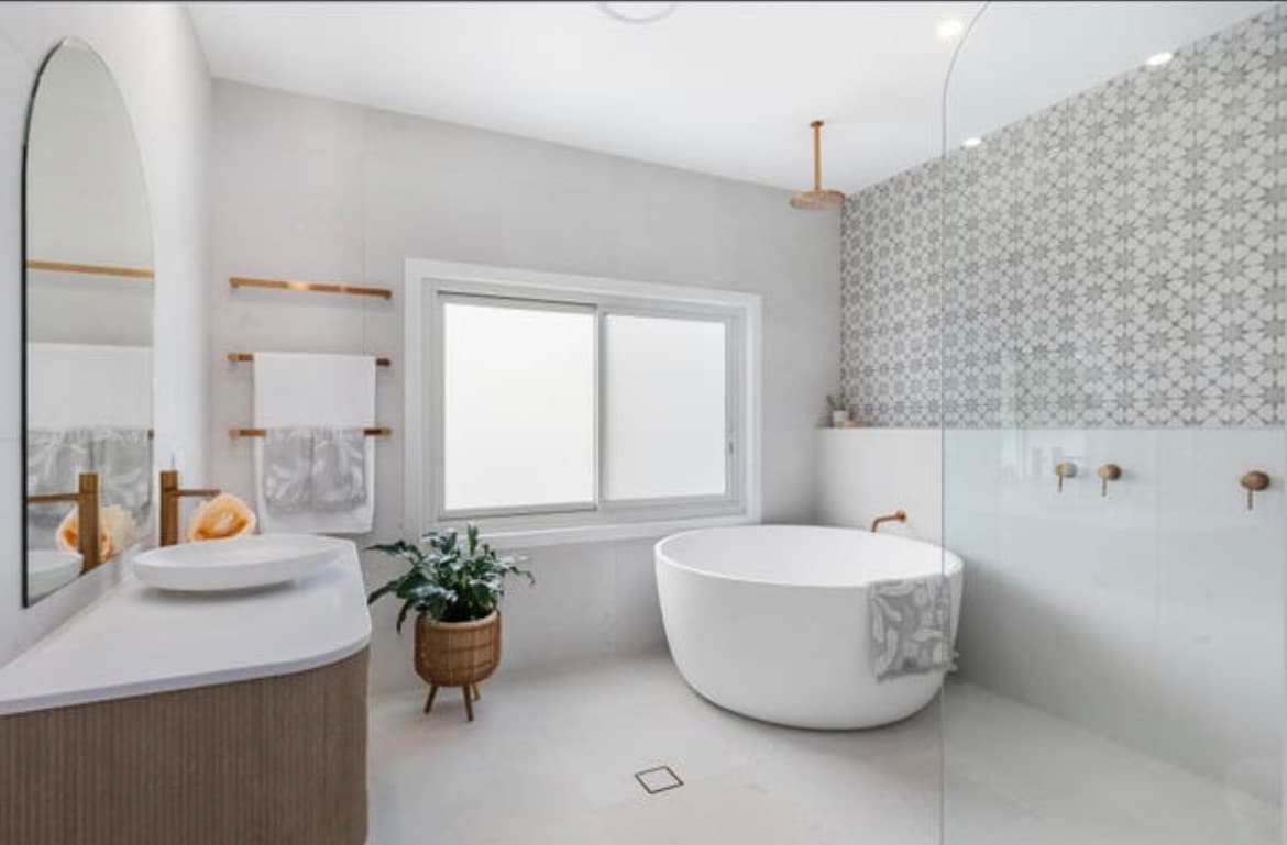 A modern bathroom features a freestanding round bathtub and a spacious glass shower. Natural light fills the room through a large window, complemented by a decorative tiled wall. Simple shelves hold neatly folded towels, while a potted plant adds a touch of greenery.