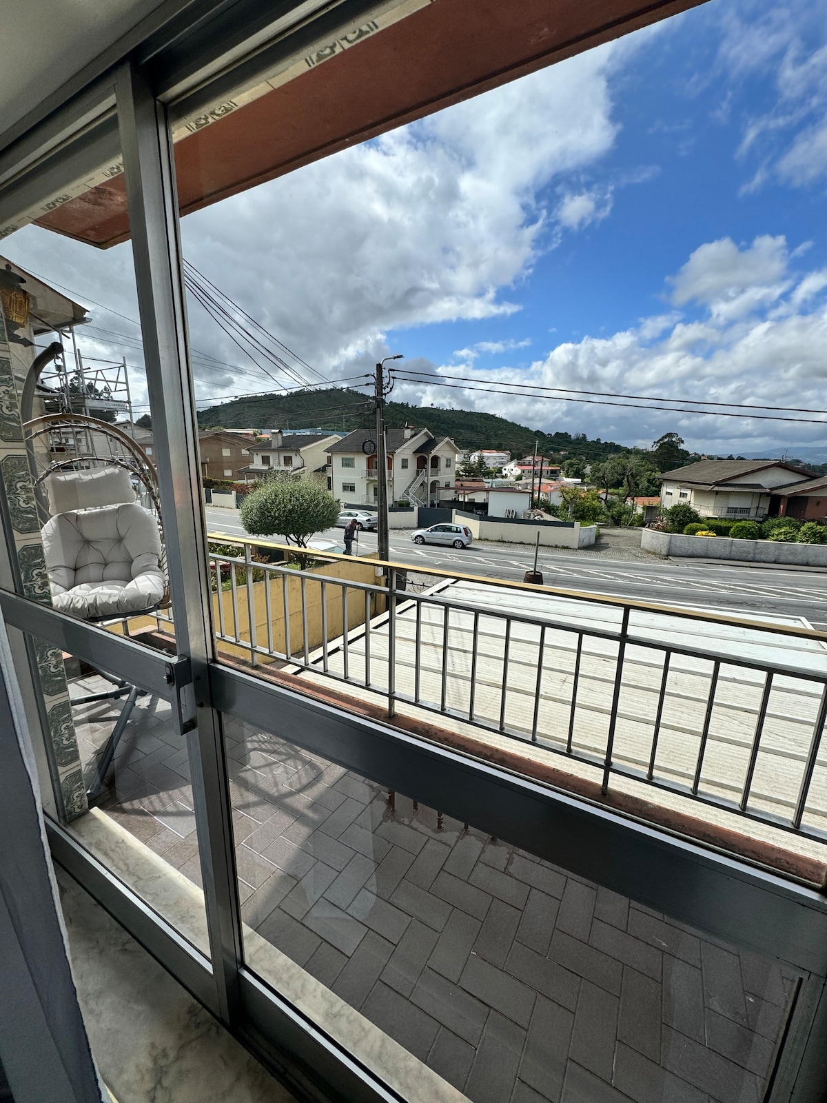 A spacious balcony is visible, showcasing views of nearby houses and a mountainous backdrop. Natural light fills the area through large glass doors, enhancing the open feel. The setting offers a glimpse of the street below, allowing for a sense of connectivity with the surroundings.