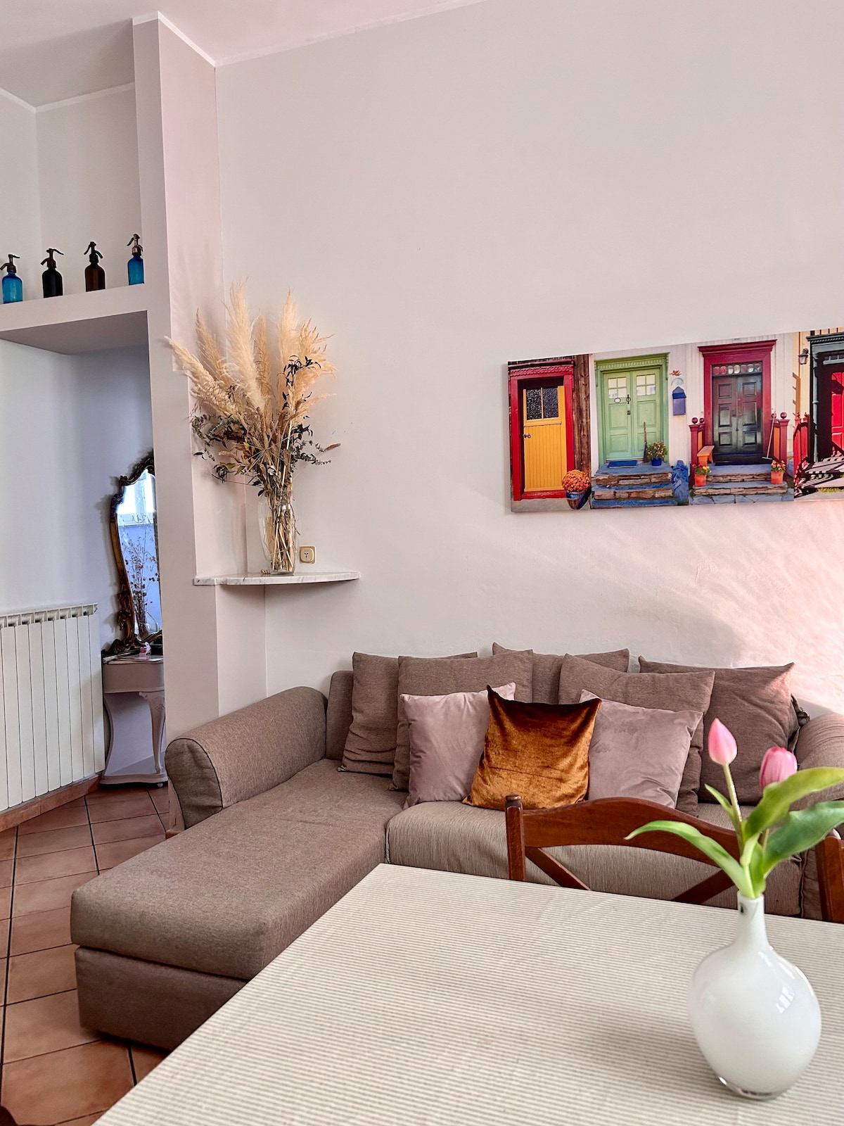 A cozy living area is depicted, featuring a light-colored sectional sofa adorned with colorful pillows. A dining table is positioned nearby, accompanied by a vase of fresh tulips. A vibrant wall artwork displaying doors enhances the cheerful ambiance, while decorative elements are placed on the shelf above.