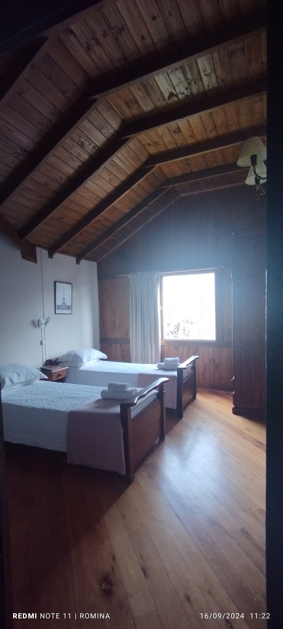 A room is furnished with two beds positioned parallel to each other, featuring wooden frames and light-colored linens. Natural light enters through a window adorned with sheer curtains, illuminating the wooden ceiling and floors. A small bedside lamp and a framed picture add to the decor.