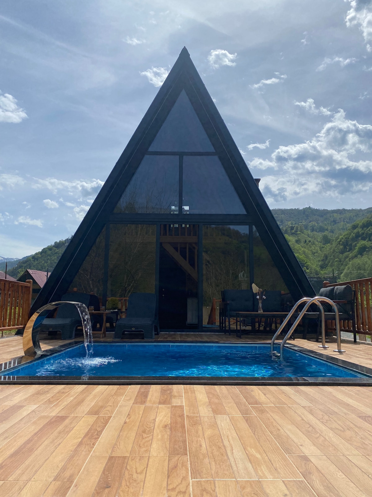An A-frame cabin features large glass windows that allow natural light to fill the interior. In front, a small pool is visible, with gentle water features and a wooden deck surrounding it. Lounge chairs are positioned nearby, offering a space for relaxation.