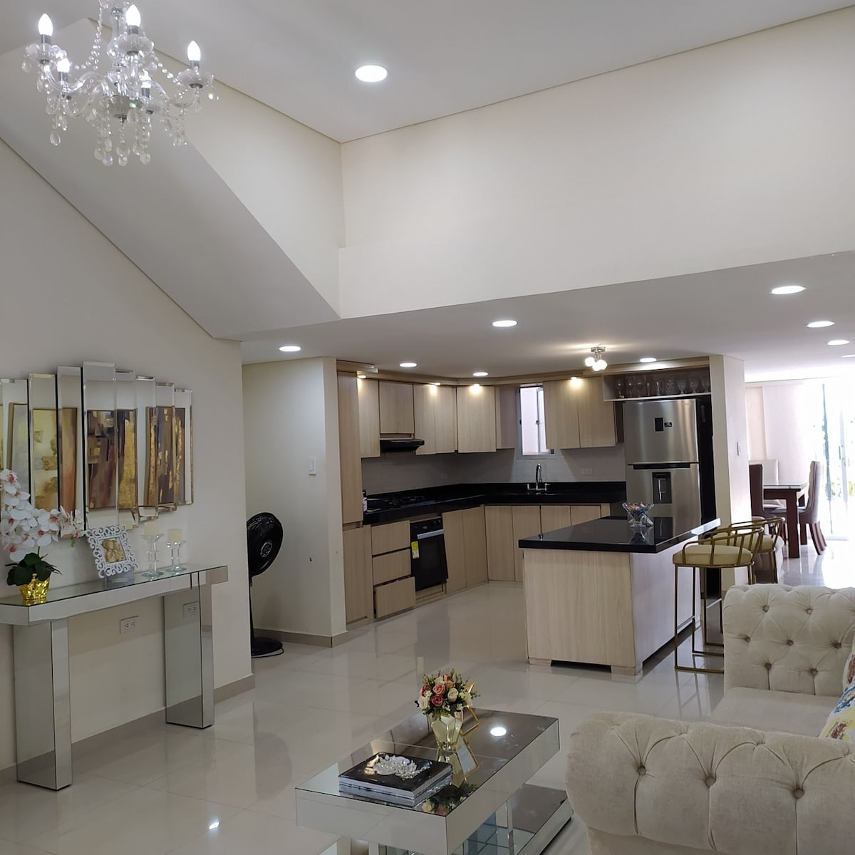An open-concept living space features a modern kitchen with sleek cabinetry and stainless steel appliances. A chandelier hangs from the ceiling, illuminating the area. A glass-topped coffee table rests on a tiled floor, complemented by a plush sofa and a decorative console table against the wall.