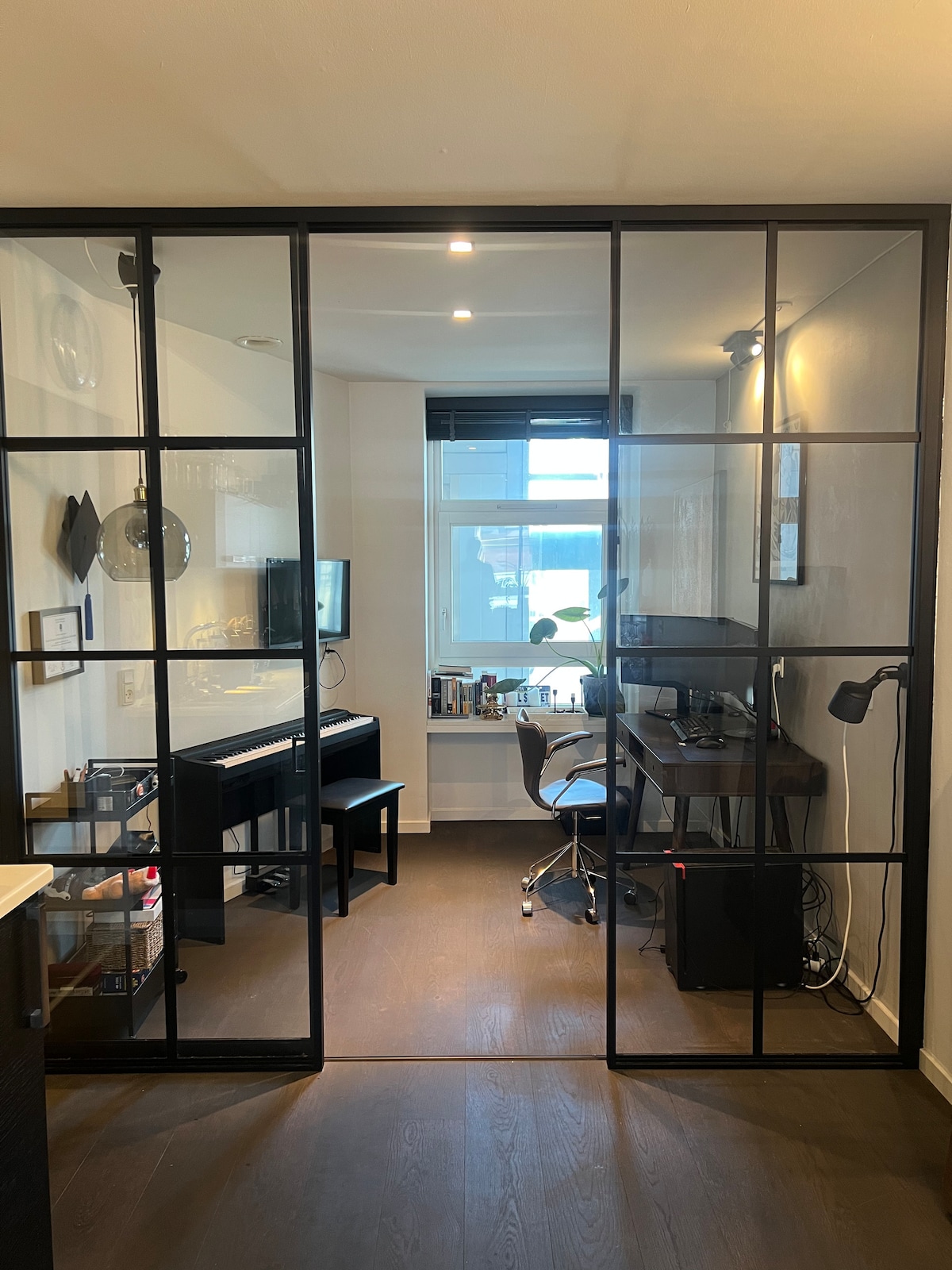 A modern workspace is visible through glass-paneled walls, featuring a desk with a computer setup and a comfortable chair. A piano and shelving with books and decorative items are also present. Natural light enters from a nearby window, enhancing the workspace ambiance.