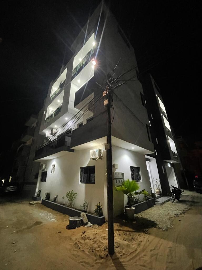 The exterior of a modern four-story building is illuminated at night, showcasing clean lines and minimalistic design. The façade features several balconies and large windows. Surrounding greenery includes potted plants along the ground level, contributing to a welcoming atmosphere.
