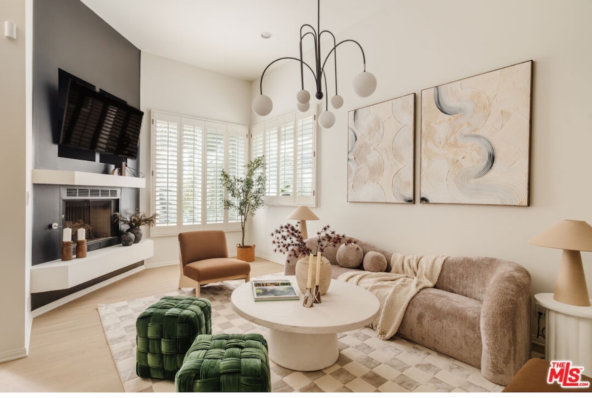 The living room features a modern design with a curved sofa, accent chairs, and a round coffee table. Natural light filters through plantation shutters, illuminating the artwork on the walls. A fireplace adds a cozy touch to the space, complemented by decorative plants and soft textiles.