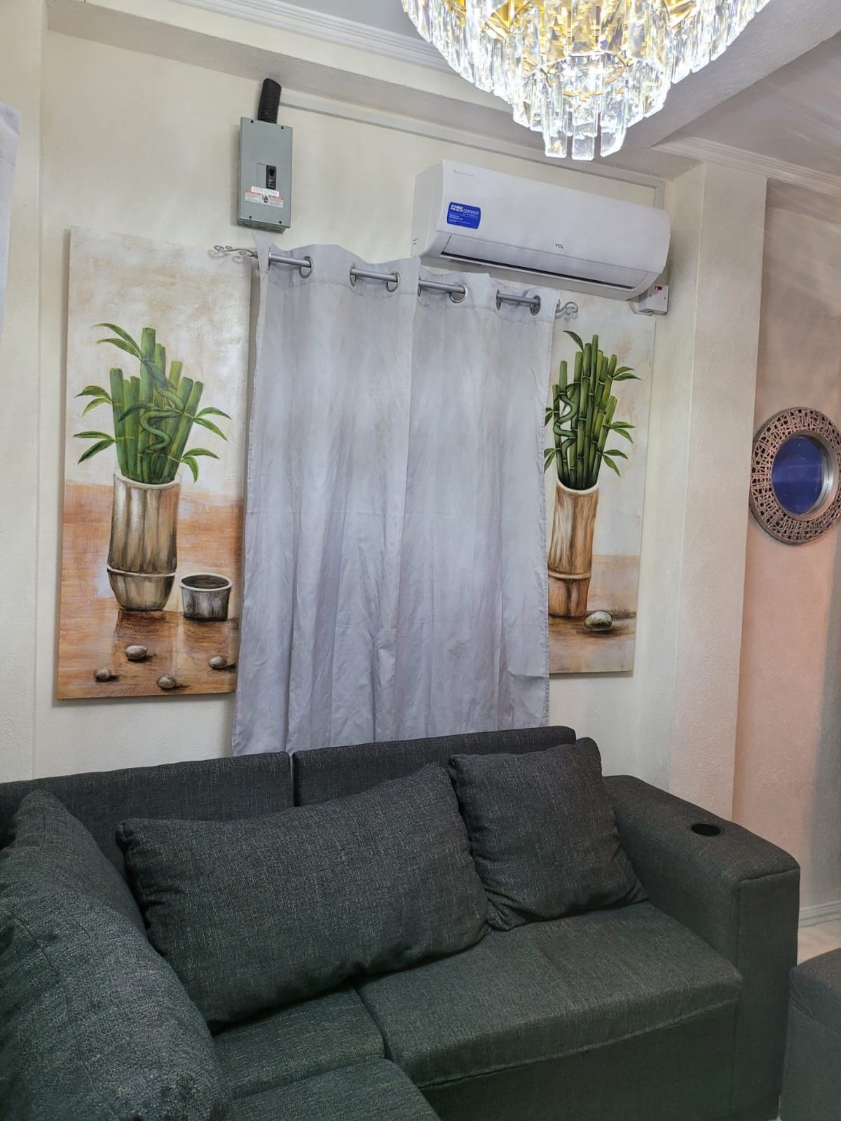 A comfortable seating area features a dark grey sectional sofa complemented by two decorative paintings of bamboo in vases. Natural light is filtered through sheer curtains, and an air conditioning unit is visible above, providing climate control. A round mirror adds a reflective detail to the space.