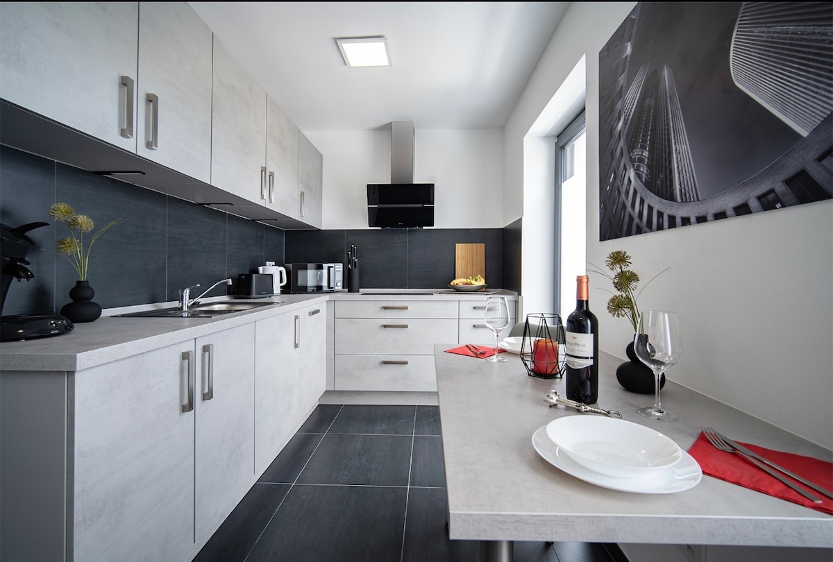 A modern kitchen features sleek cabinetry with a minimalist design. Countertops display neatly arranged table settings, including plates and glasses. A dining table is adjacent, accompanied by decorative elements. Natural light enters through a nearby window, highlighting the clean lines and contemporary appliances.