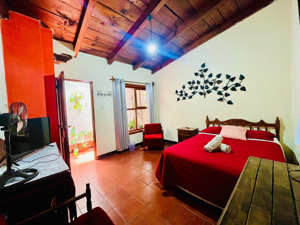 A cozy bedroom features a spacious bed with a red coverlet, complemented by decorative pillows. Natural light is allowed in through a window with sheer curtains, while a wall adorned with leaf motifs adds an artistic touch. A wooden table and two red chairs provide additional seating.