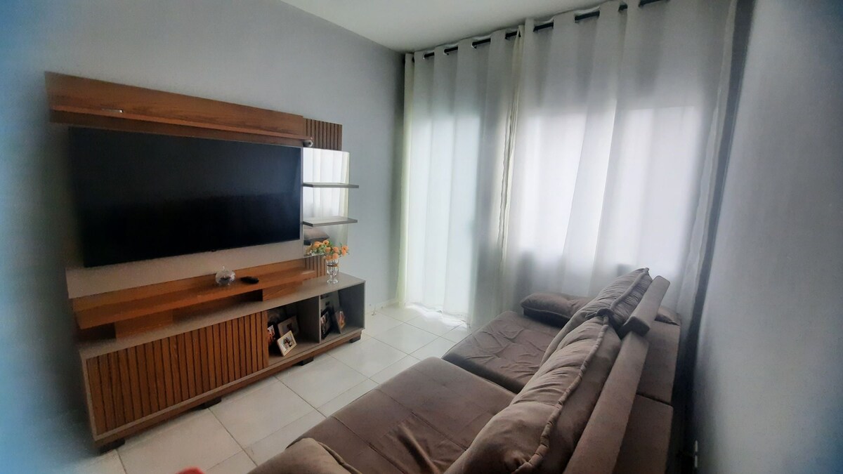 A comfortable living area is showcased, featuring a large television mounted on a wooden console. A sectional sofa with soft cushions is positioned nearby, complemented by sheer curtains that allow natural light to filter in through the windows.