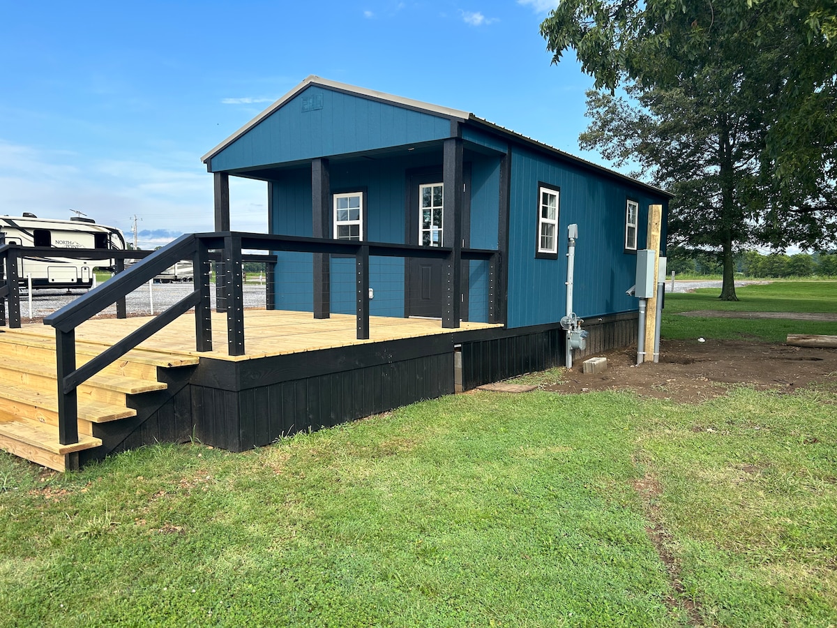 A newly constructed tiny house is showcased with a modern blue exterior. A spacious wooden front deck offers access via steps, inviting relaxation outdoors. Surrounding grassy areas are visible, enhancing the property’s natural setting. The house features two windows allowing light into the interior.