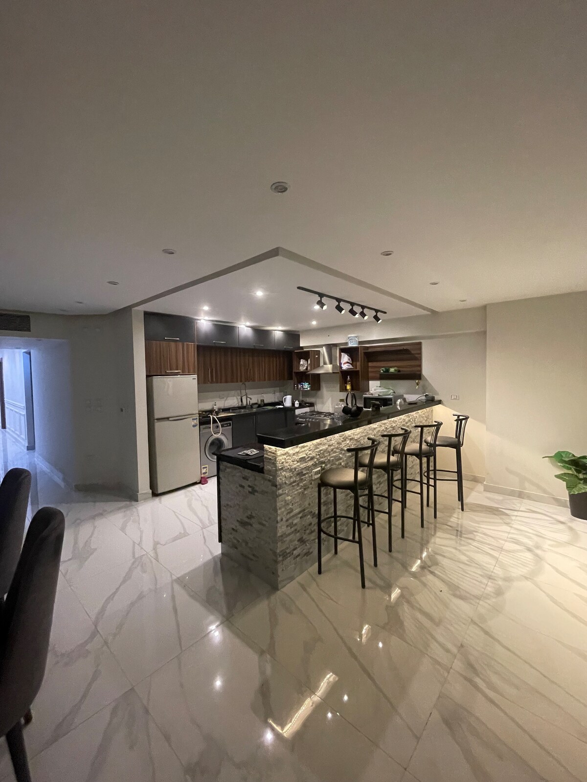 A modern kitchen is shown, featuring a spacious island with bar seating. Sleek cabinetry and stainless steel appliances are complemented by polished marble flooring. Soft lighting enhances the contemporary design, creating a functional cooking space.