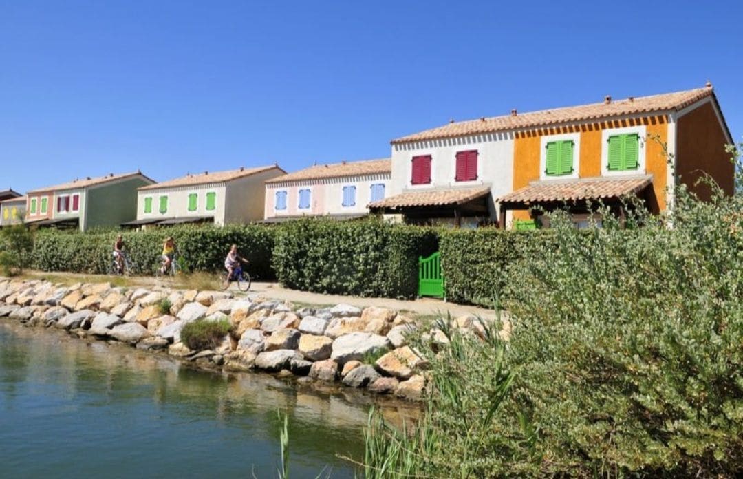 A row of colorful two-story homes is lined along a serene waterway. Each house features vibrant façades with green shutters and terracotta roofs. A path runs alongside the water, with cyclists enjoying the tranquil surroundings, framed by lush greenery and smooth stones by the water's edge.