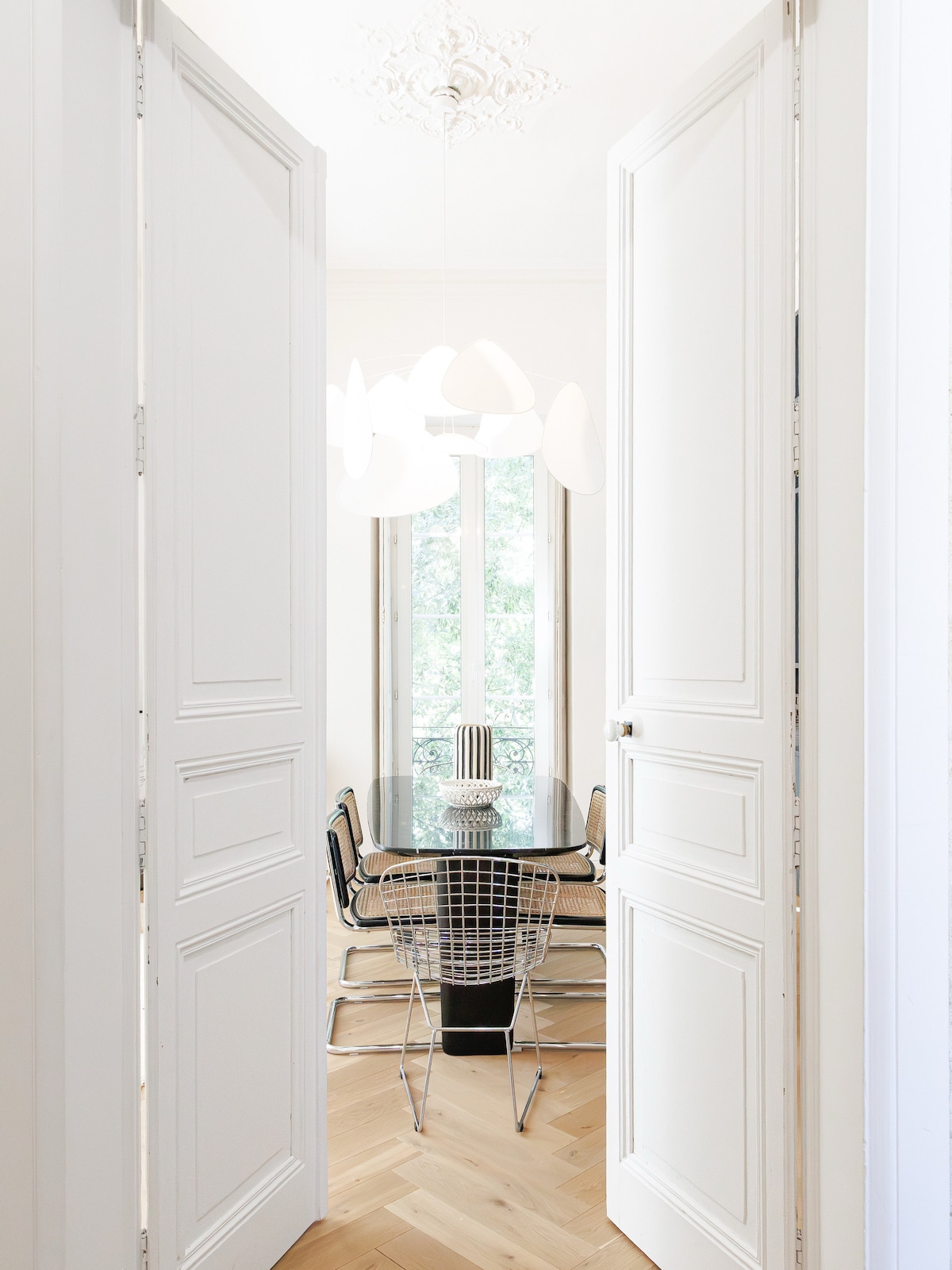 A bright dining area is visible through open double doors, featuring a modern glass dining table surrounded by sleek chairs. Large windows allow natural light to fill the space, highlighting the stylish decor and creating a welcoming atmosphere.