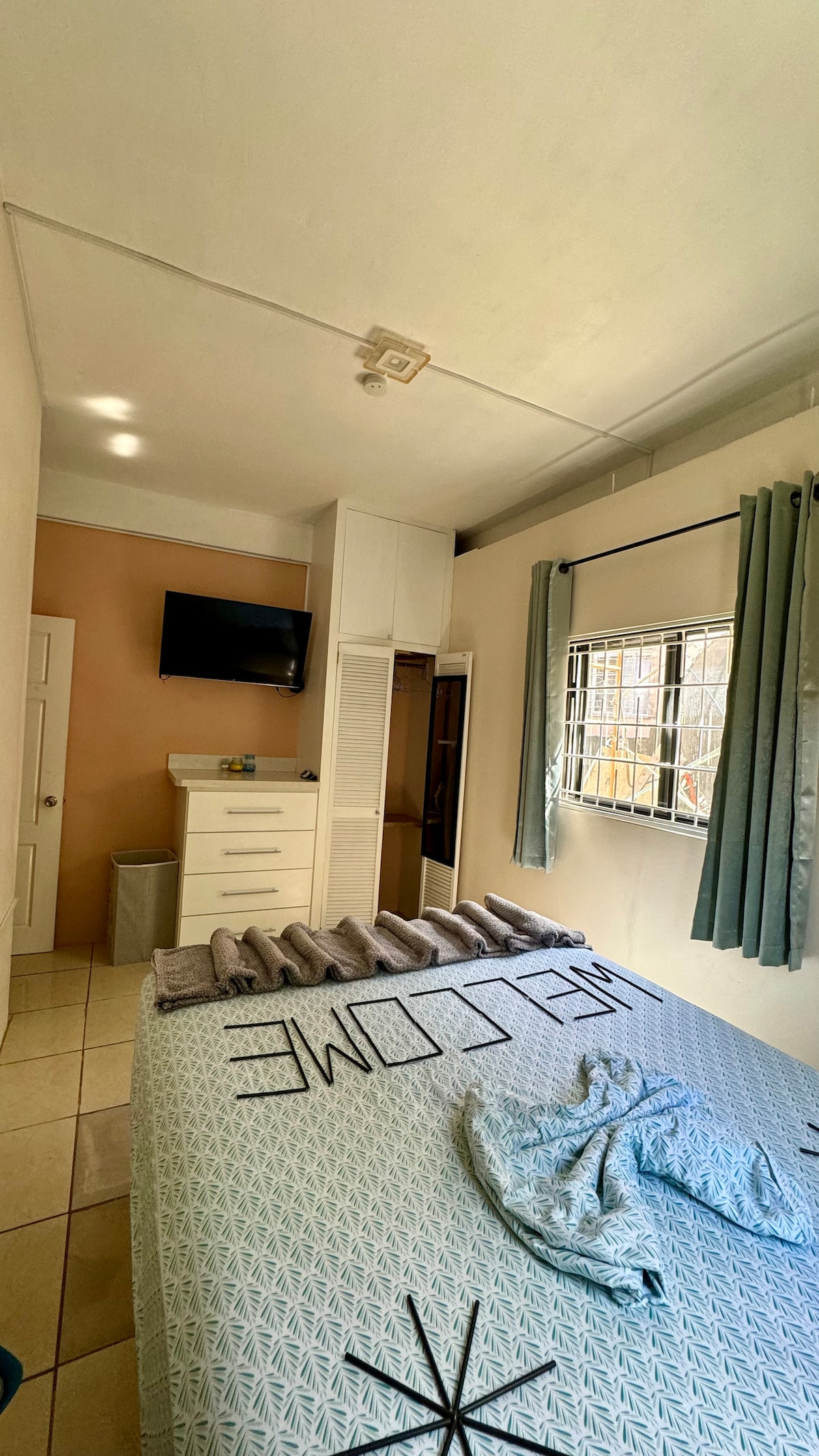 A well-lit bedroom is displayed, featuring a spacious bed with decorative bedding. A flat-screen TV is mounted on the wall opposite the bed. A wardrobe and dresser are positioned nearby, and a window is adorned with light green curtains, allowing natural light to fill the space.