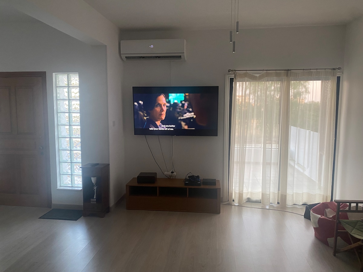 A spacious living room features a wall-mounted television displaying content. Natural light filters through sheer curtains, enhancing the airiness of the space. A wooden cabinet stands beneath the TV, while a doorway with a glass insert introduces the outdoors.