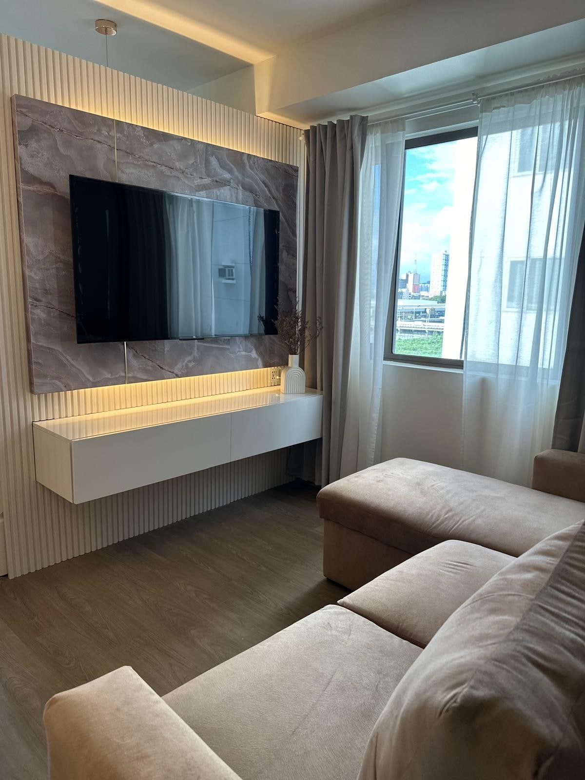 The living area features a sleek, wall-mounted television above a modern white console. A cozy sofa in a light tone provides ample seating. Natural light filters through a large window with sheer curtains, offering views of the city skyline in the background.