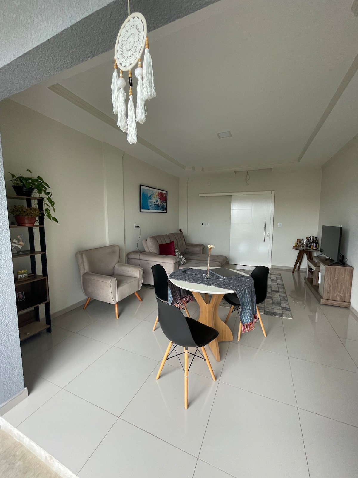 A shared living space features a round wooden dining table surrounded by four black chairs. A comfortable gray sofa is positioned nearby, accompanied by a neutral rug. Natural light floods the area through a nearby window, with a plant accentuating the corner.