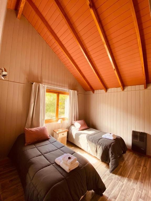 A cozy room features two spacious beds with dark bedding, each adorned with folded white towels. Natural light filters in through a window, enhancing the wooden ceiling and walls with warm tones. A small bedside table is placed beside one of the beds.
