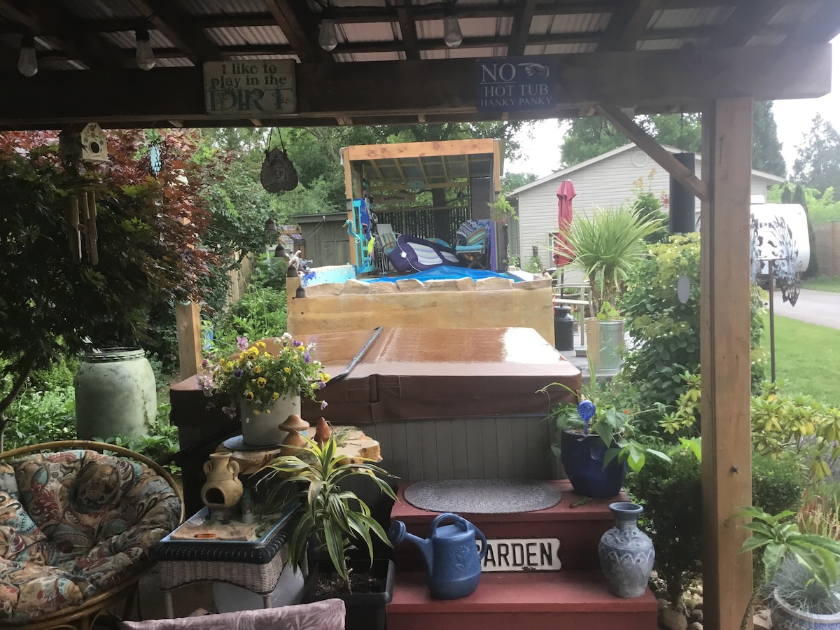 A covered outdoor space is framed by greenery, featuring comfortable seating areas with floral cushions. A wooden deck leads to a hot tub, surrounded by plants. A sign reading 'GARDEN' is visible on the steps, enhancing the inviting atmosphere.