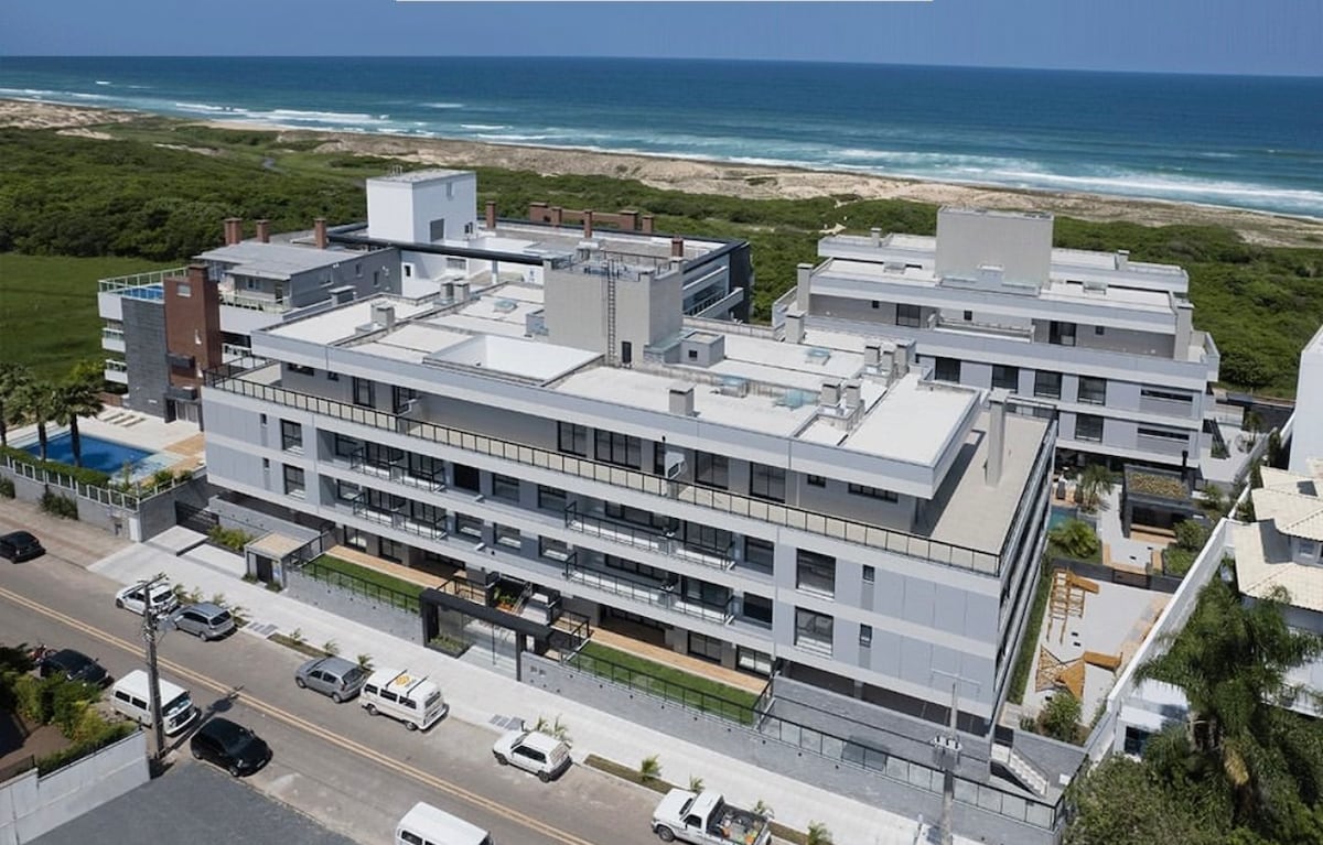 An aerial view captures the modern condominium complex situated near a beach, with multiple buildings outlined against the coastline. The sparkling ocean reflects the sunlight, and green spaces surround the property, conveying a serene atmosphere. A pool is visible in the foreground.