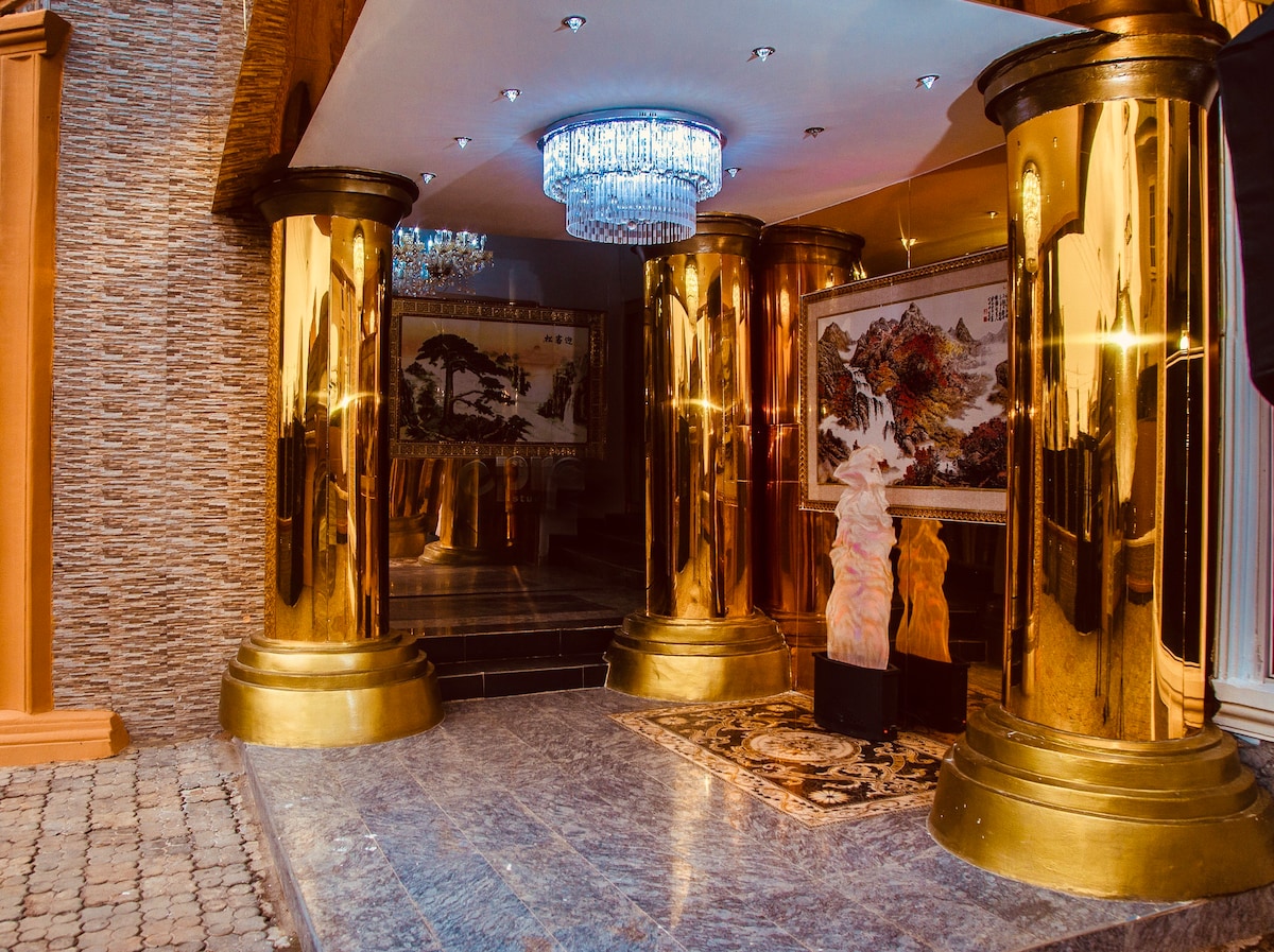 The entrance area features striking golden columns, which reflect light from an elegant chandelier above. A decorative rug adds texture to the polished floor, while artwork decorates the wall, contributing to the sophisticated ambiance of the space.