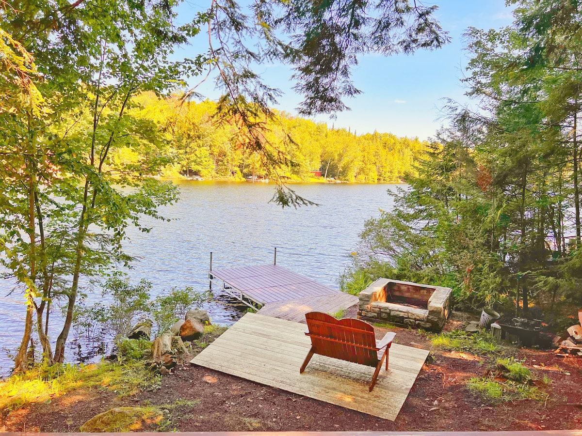The lakeside area features a wooden dock extending into the calm waters of Lake Tekeni. A rustic fire pit is visible on a deck made of wooden planks, surrounded by lush greenery and rocks, providing a serene spot to relax amidst nature.