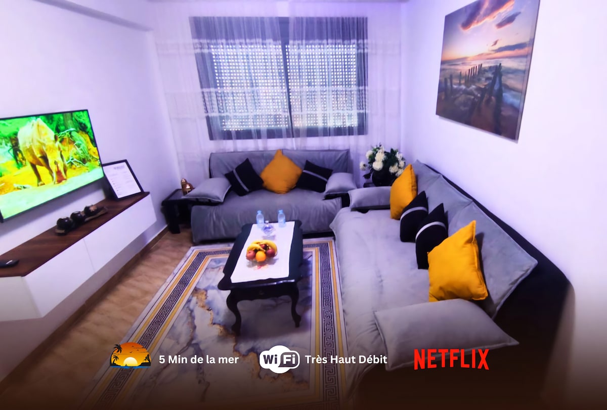 A cozy living area features a large sectional sofa adorned with orange and black accent pillows. A coffee table sits at the center, complemented by a decorative rug. A wall-mounted TV displays vivid images, and natural light filters through sheer curtains.